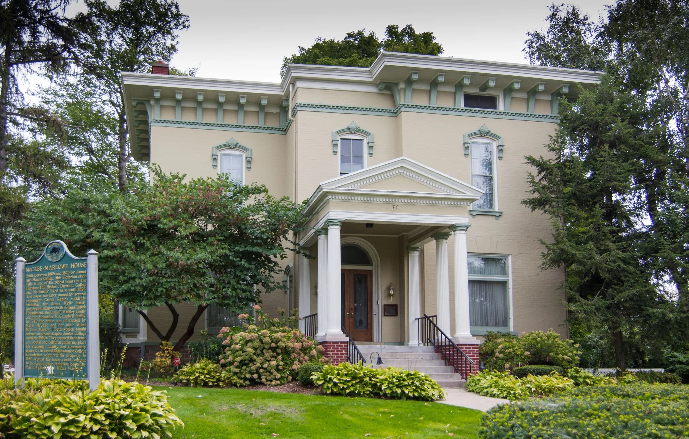 A two-storey cream-rendered house with tall arched windows, deep bracketed eaves and a small columned entrance porch reached by brick steps, framed by mature conifers with flowering shrubs and a clipped lawn in front.