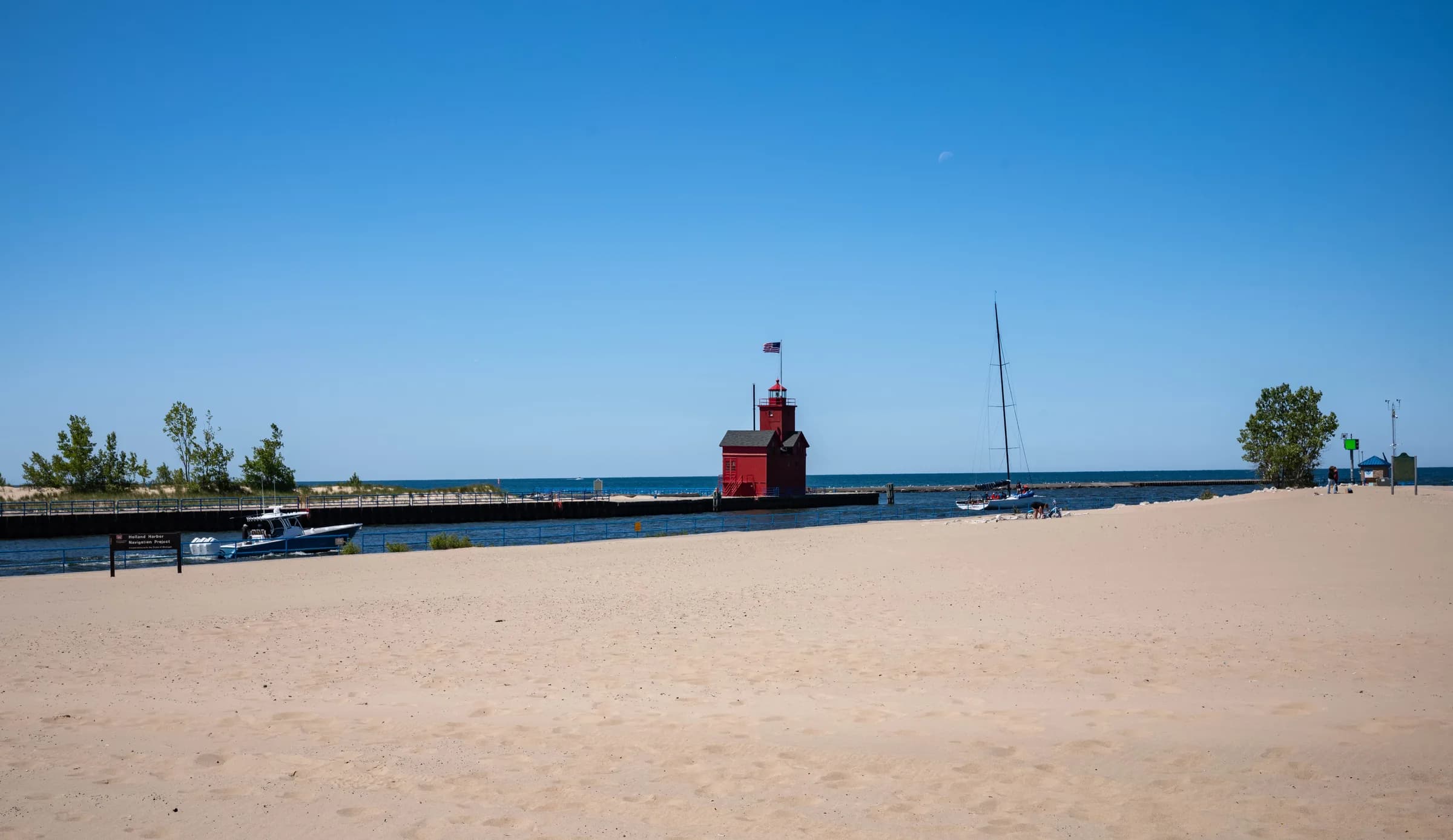A wide sweep of pale dry sand under a deep blue summer sky, with a low breakwater and pier running out to flat open water, a moored sailboat with its mast up, and a small red structure at the far end of the pier.