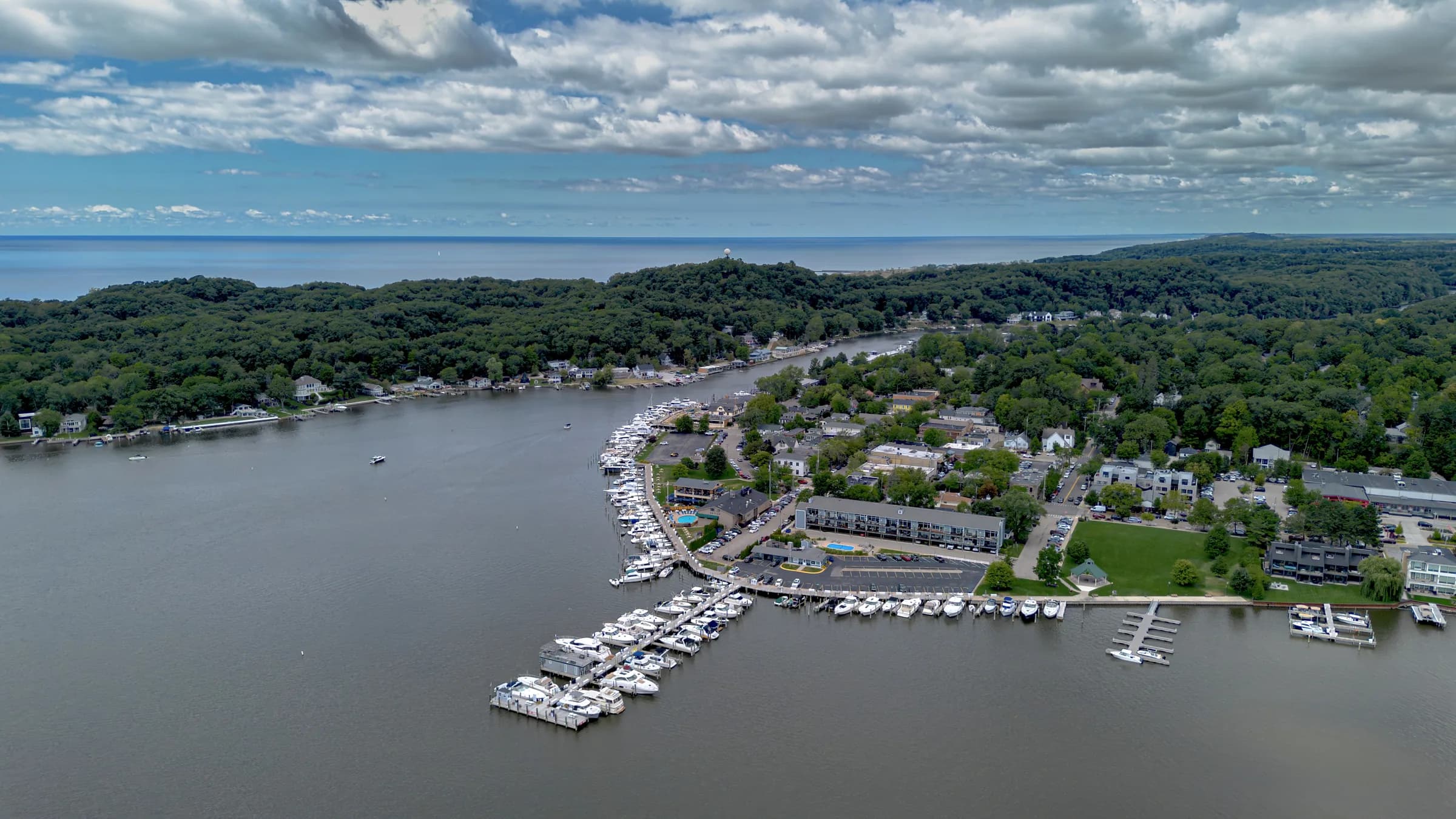 Aerial view of a small town wrapped around a wide river mouth, floating docks and moored boats filling the near bank, wooded bluffs beyond and open lake on the horizon under broken cloud.