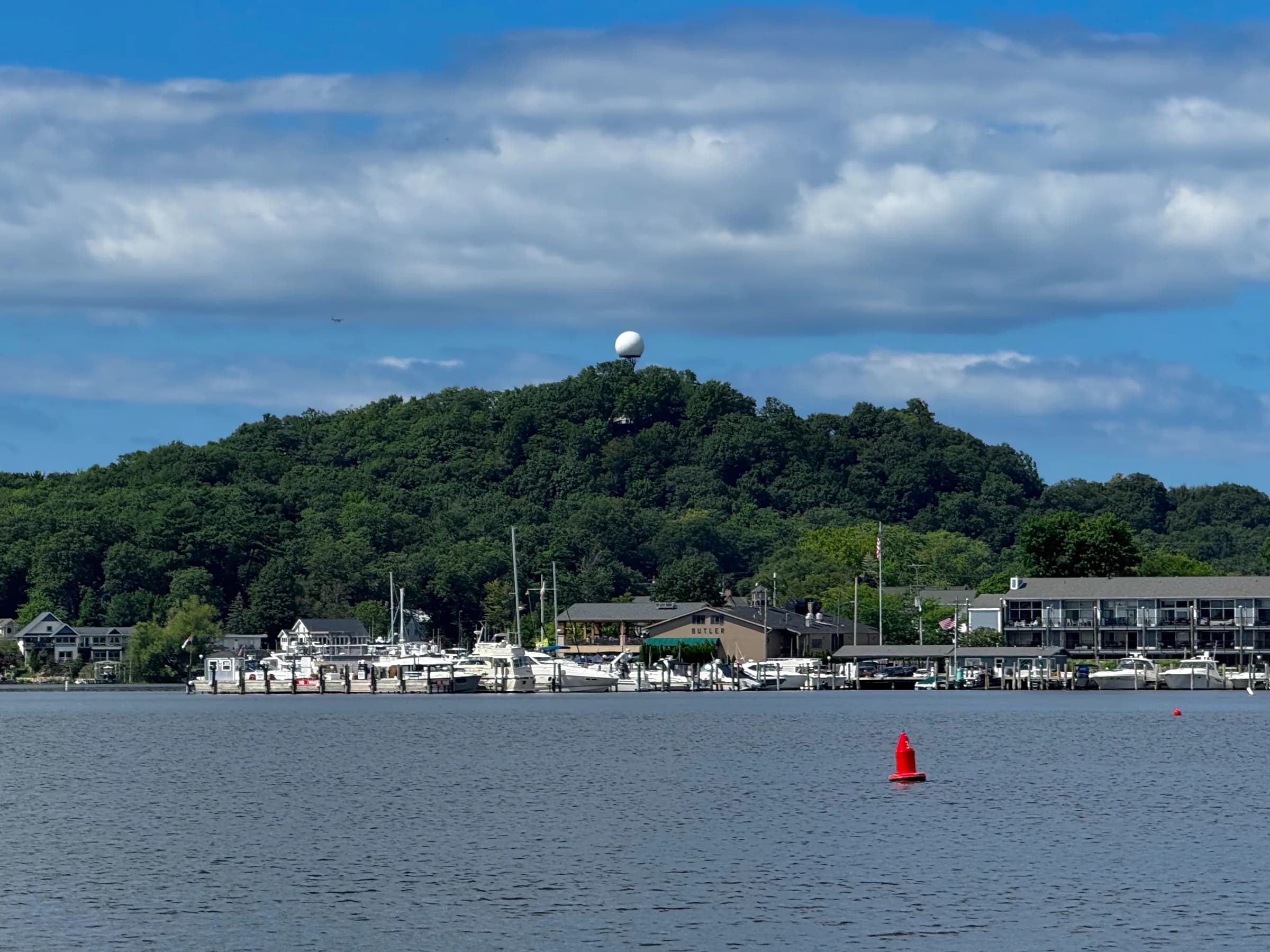 A large wooded dune rising steeply behind a line of moored boats and low waterfront buildings, seen from across grey-blue water under a sky of heavy cumulus.