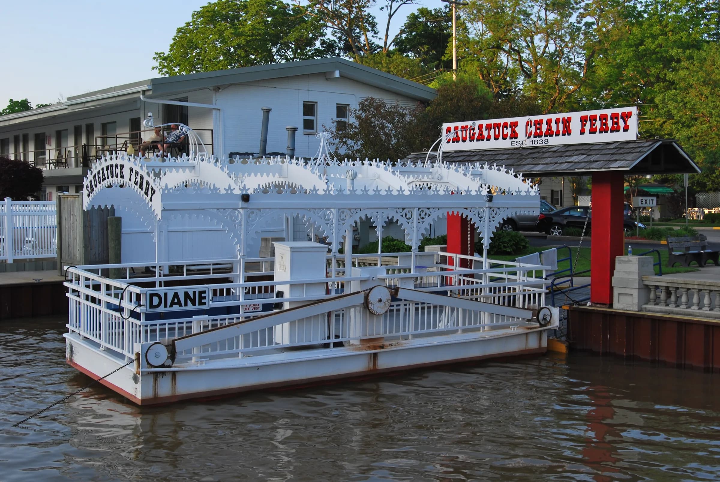 A small white passenger ferry with a decorative canopy and a red-trimmed wheelhouse moored at a dock, its name board reading Saugatuck Chain Ferry.