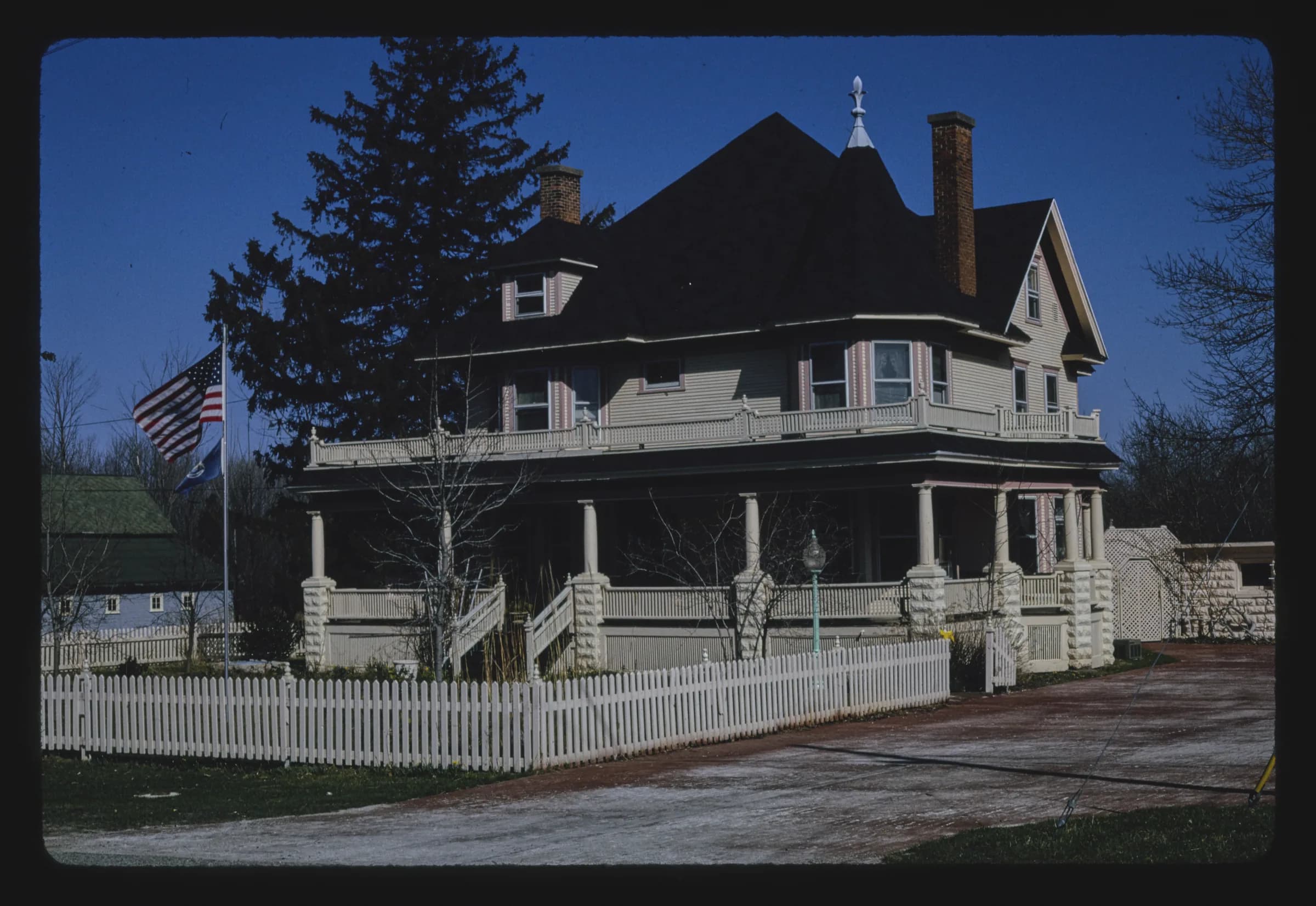 A large dark Victorian house with a corner turret, deep porch and decorative bargeboards, behind a white picket fence and a flagpole, framed by mature evergreens.