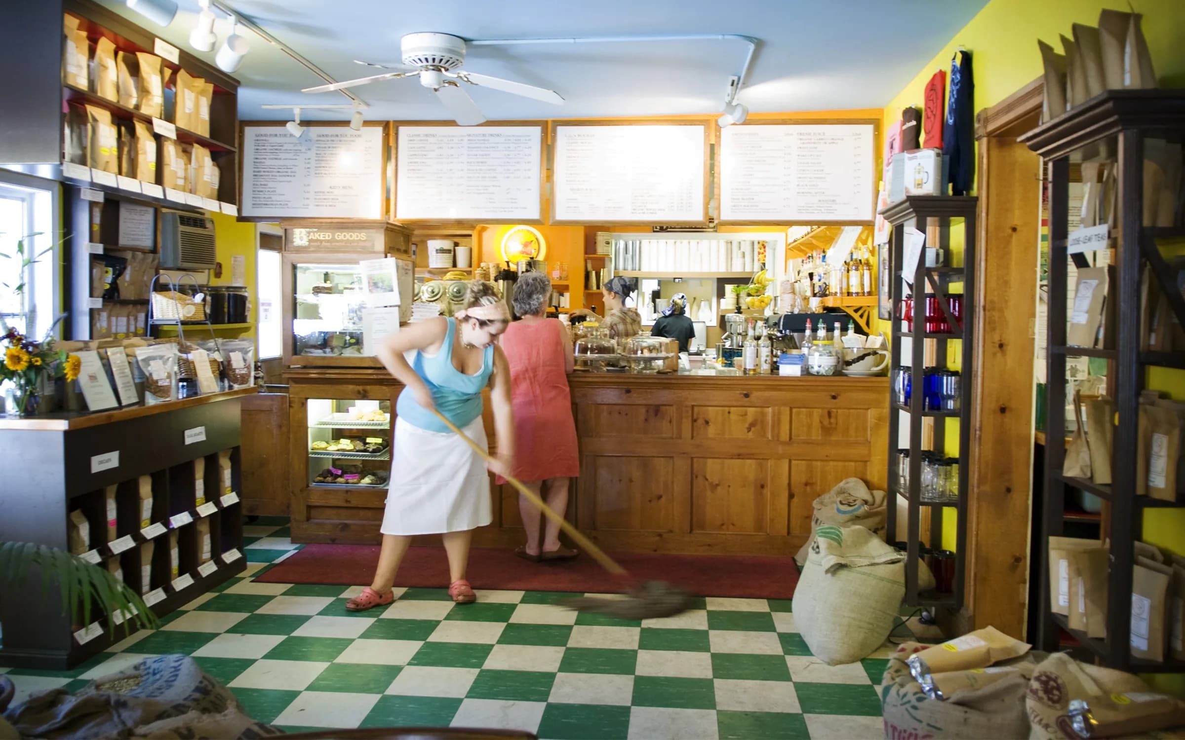 The interior of an old shop with a black and white chequerboard floor, timber counters and shelving, a menu board above and two staff working behind the counter.