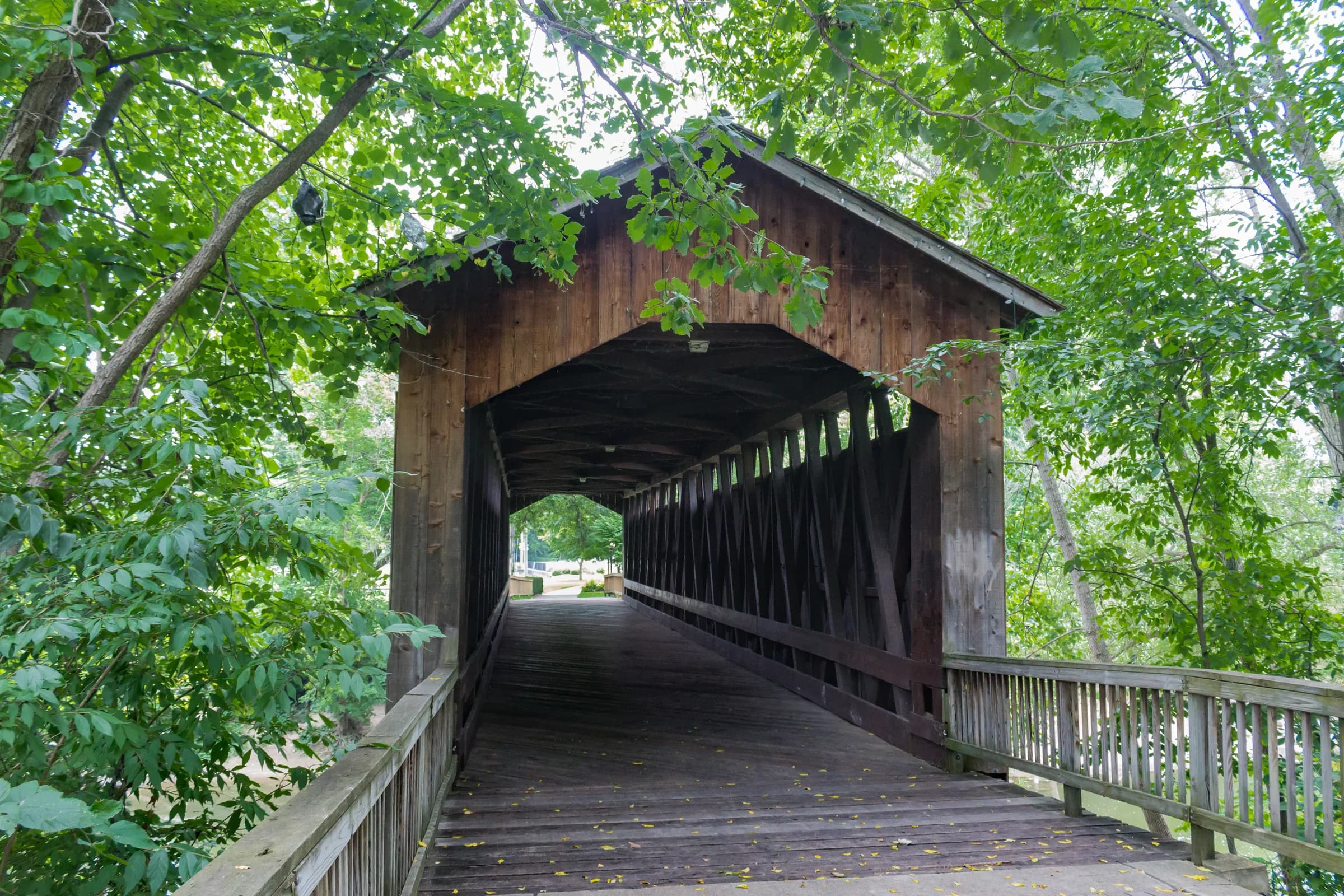 A view straight into a wooden covered bridge from its portal; the board floor and diagonal truss timbers recede toward a bright opening at the far end, with leaves hanging over the roofline.