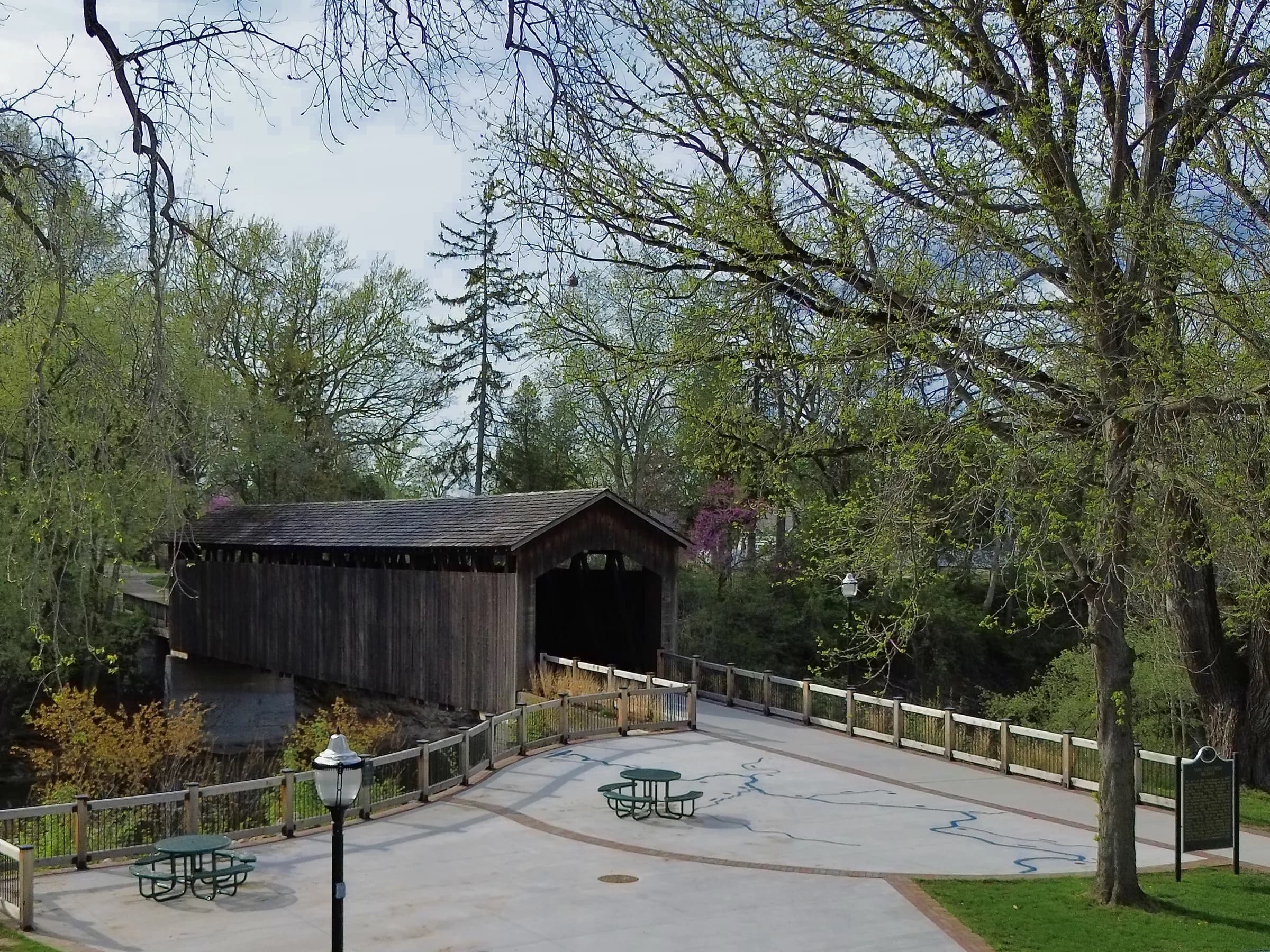 A broad concrete terrace holding picnic tables and a black lamp post curves toward a dark timber bridge, with bare and budding trees standing behind it in early spring.