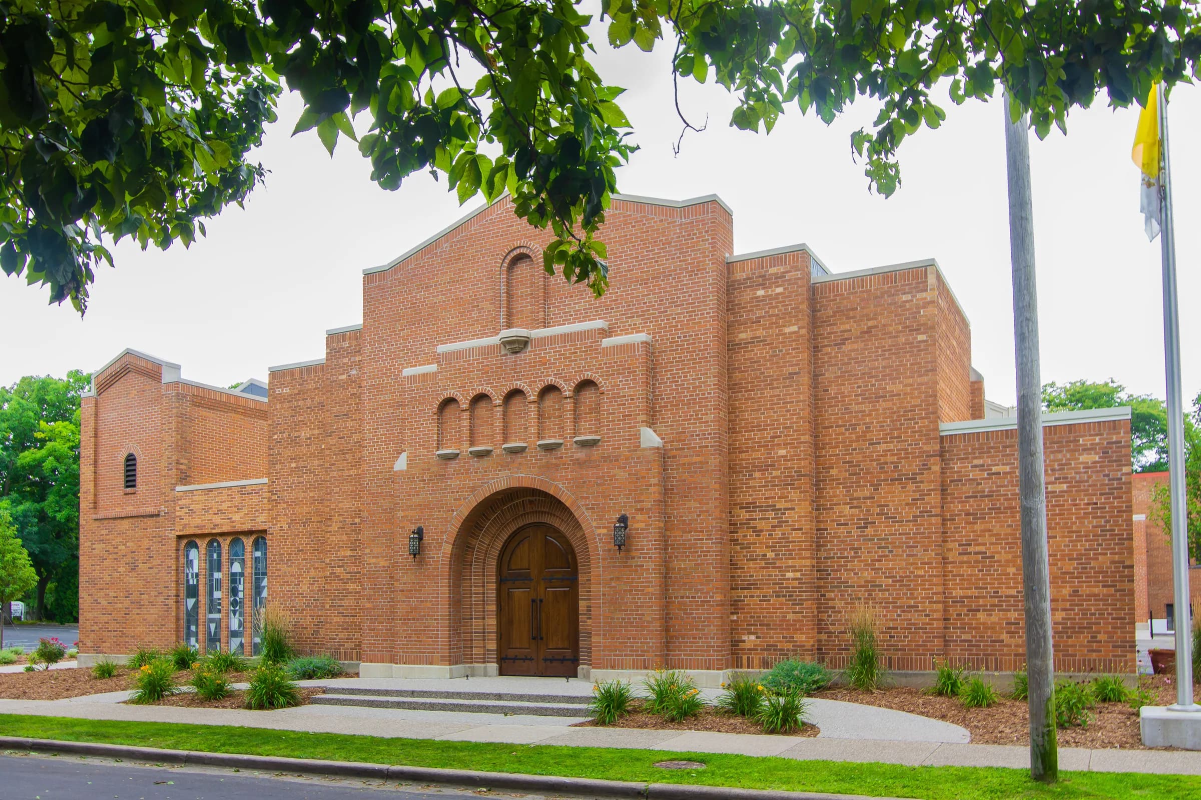 A red-brick church with a stepped gable, arcaded window band and a deep round-arched entrance with timber doors, shot in summer sun and framed overhead by green leaves.