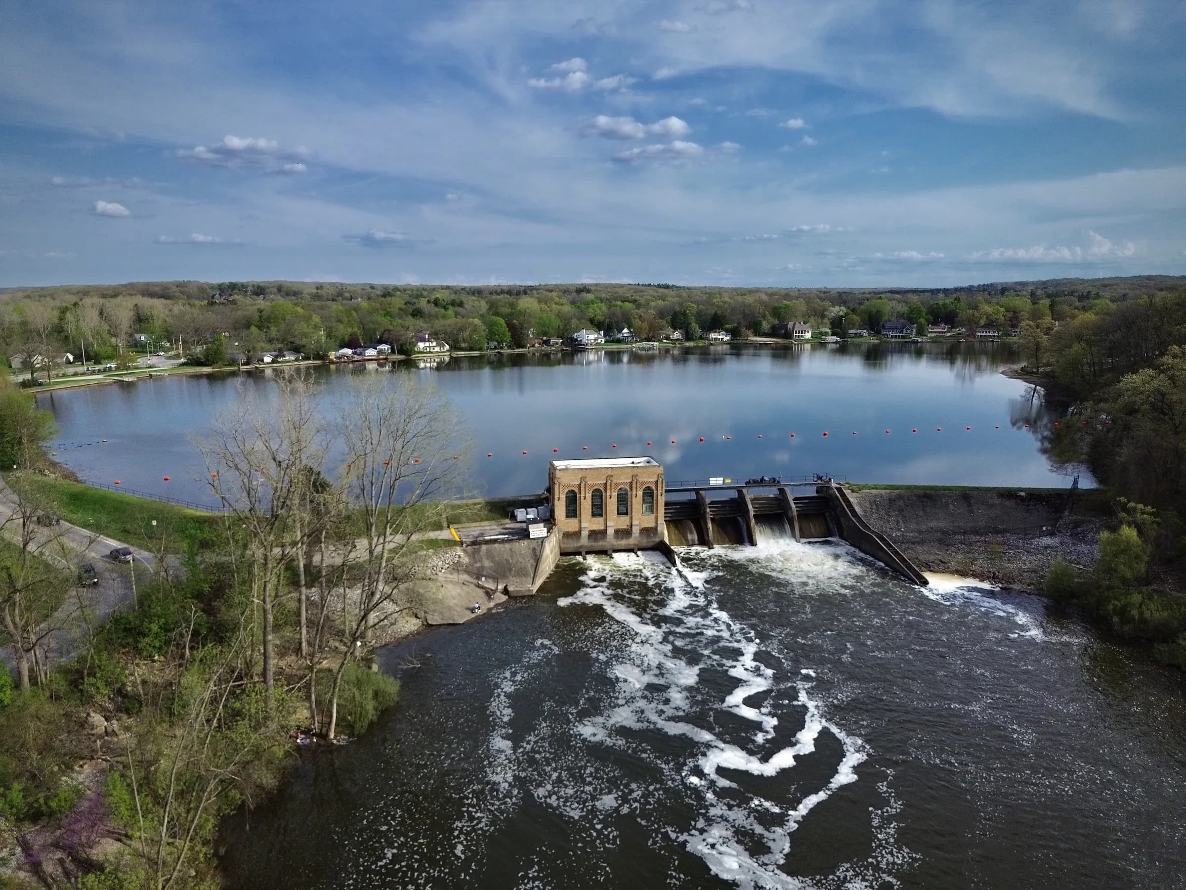 An elevated view of a low concrete dam and a small brick powerhouse; white water spills across the full width of the frame while a wide calm pond and wooded banks lie beyond under a blue sky with scattered cloud.