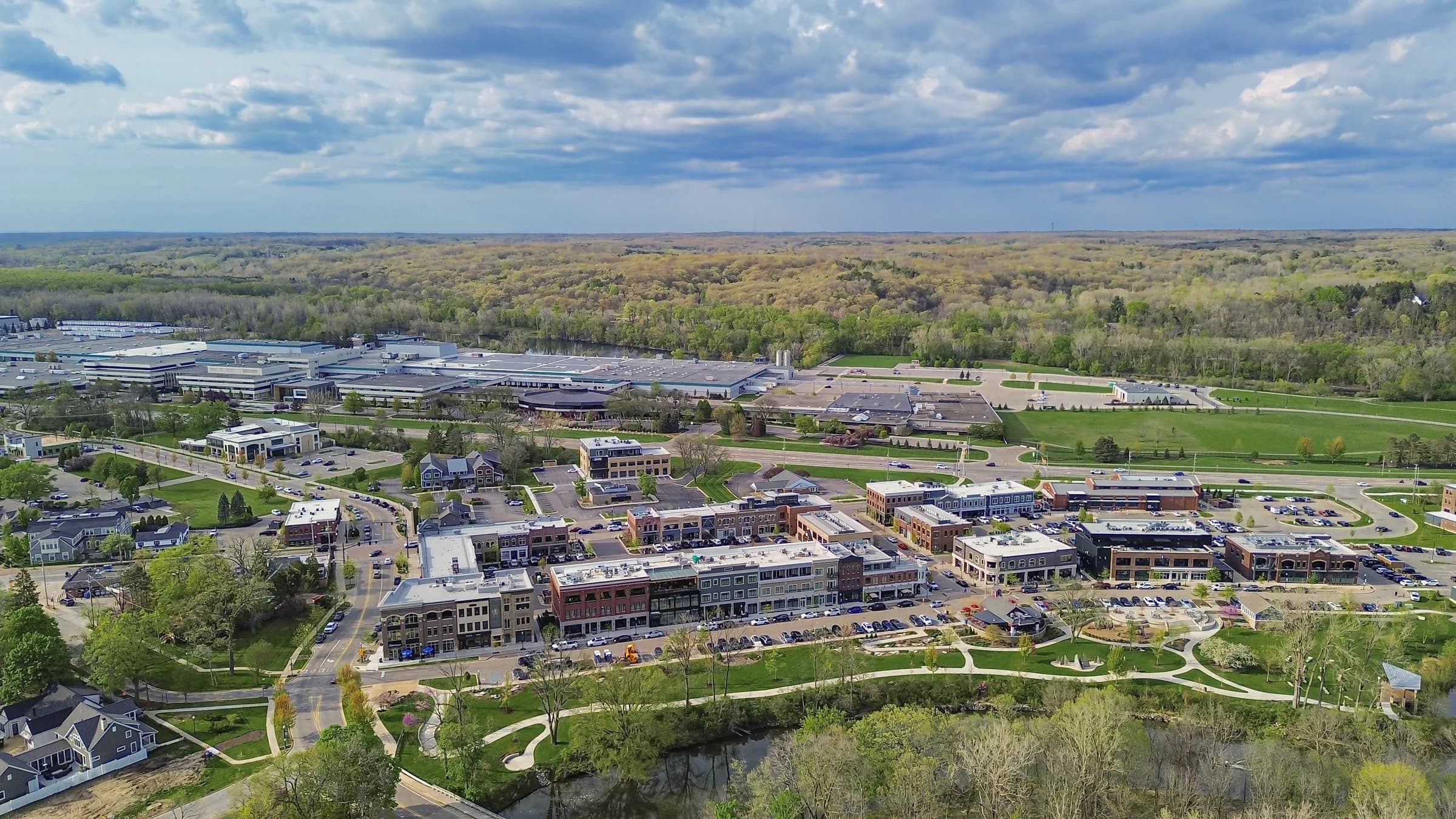 An aerial photograph of a small mixed-use town centre of low brick buildings with pitched roofs, streets and a landscaped park beside a river, with woodland and farmland running to a flat horizon under a cloudy sky.