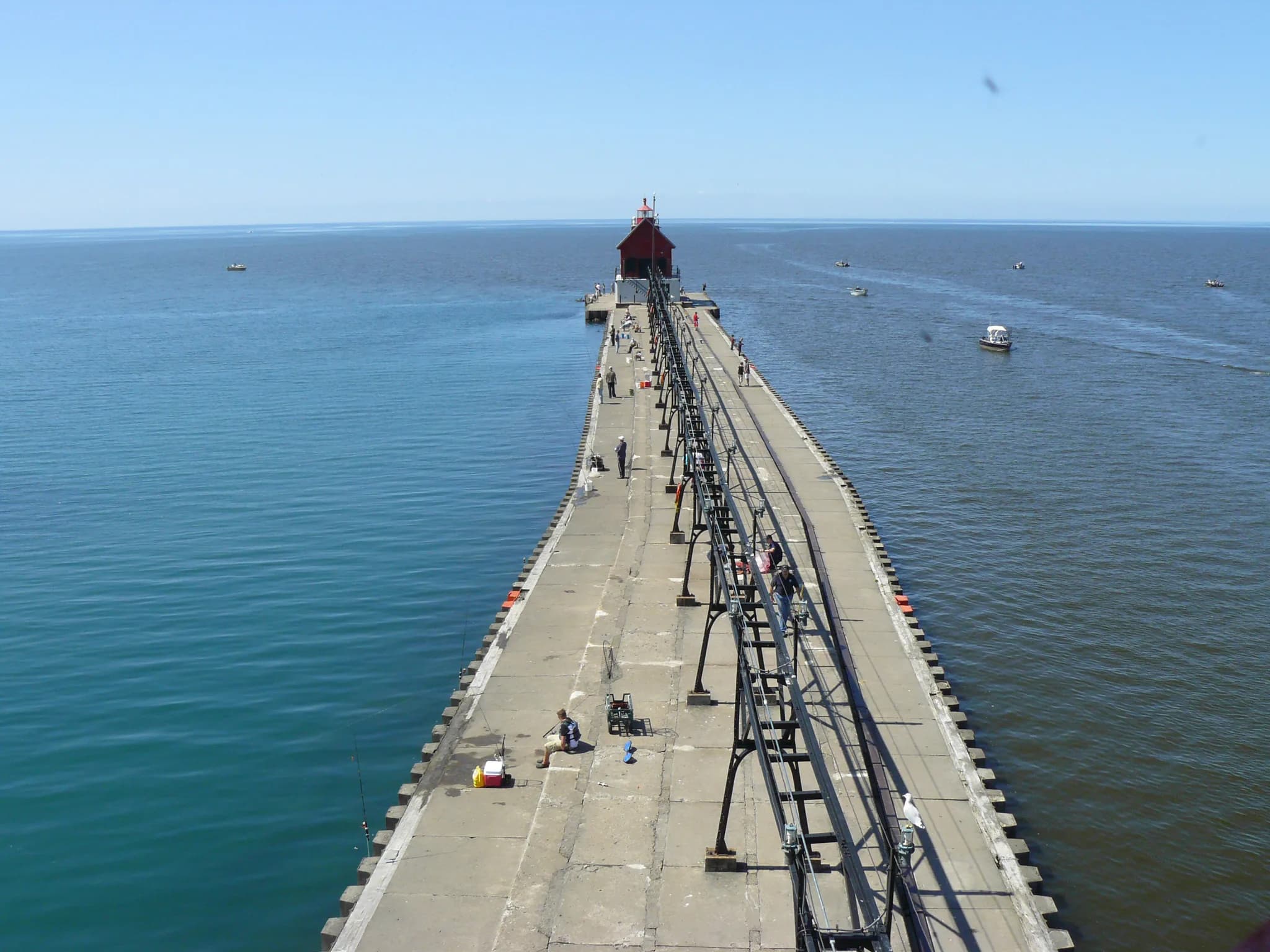 A long concrete pier with a raised steel catwalk on stilts running its full length toward a small dark lighthouse at the far end, flat blue water either side and a few people fishing from the edge.