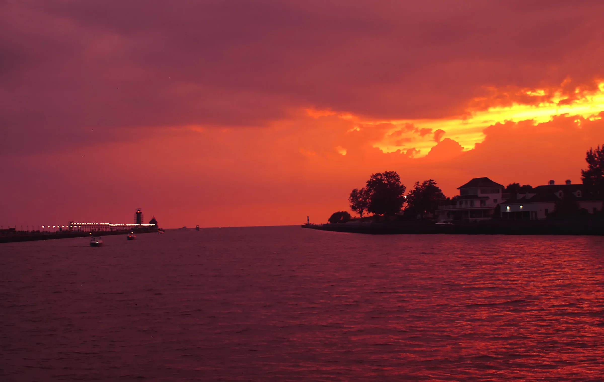 A heavy orange and red sunset burning through low cloud over dark water, the land reduced to a black silhouette along the horizon and a breakwater running in from the left.