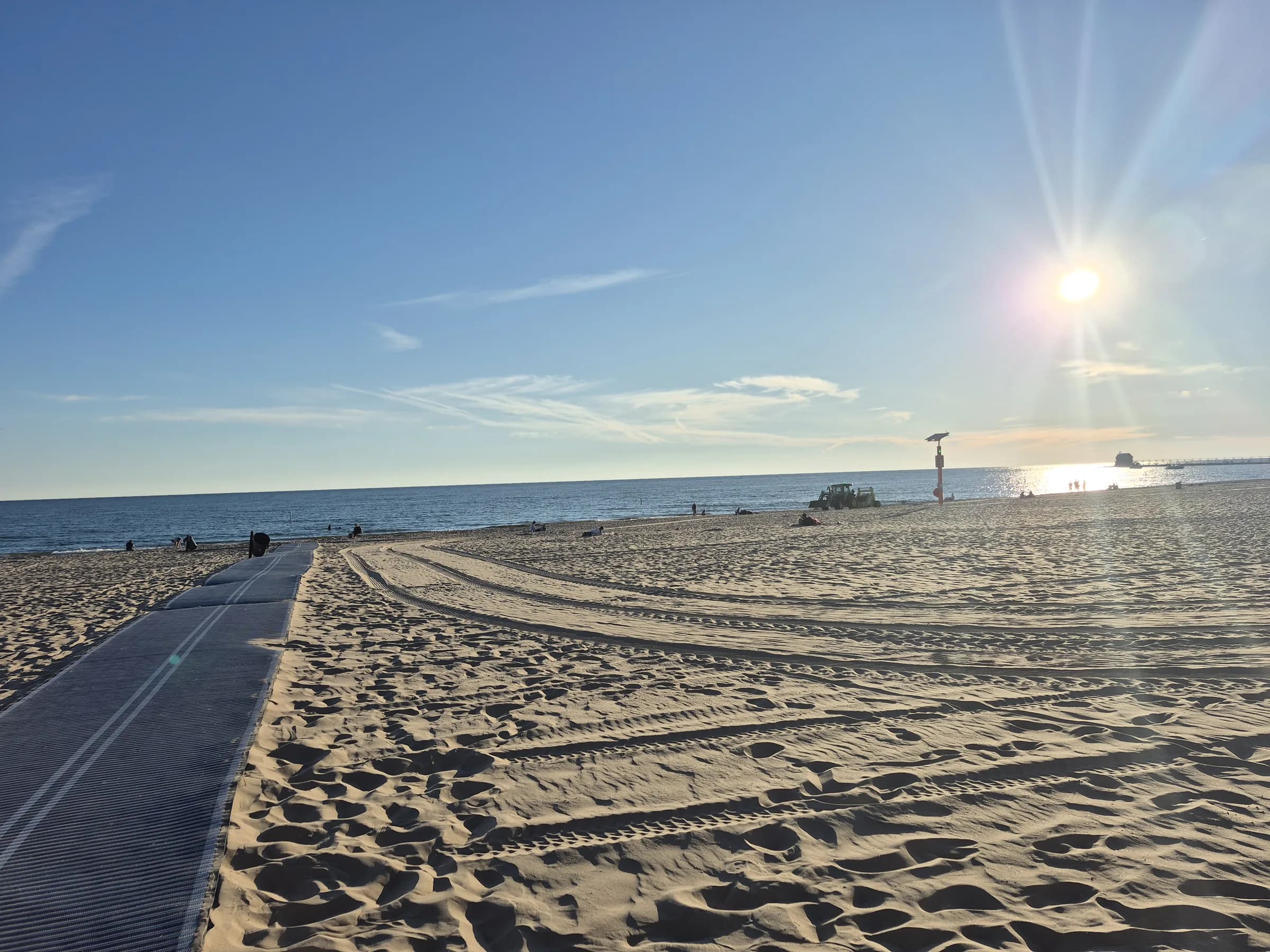 Wide raked sand in the foreground running down to flat blue water, a low pier crossing the middle distance and a bright hazy sun high over the lake.