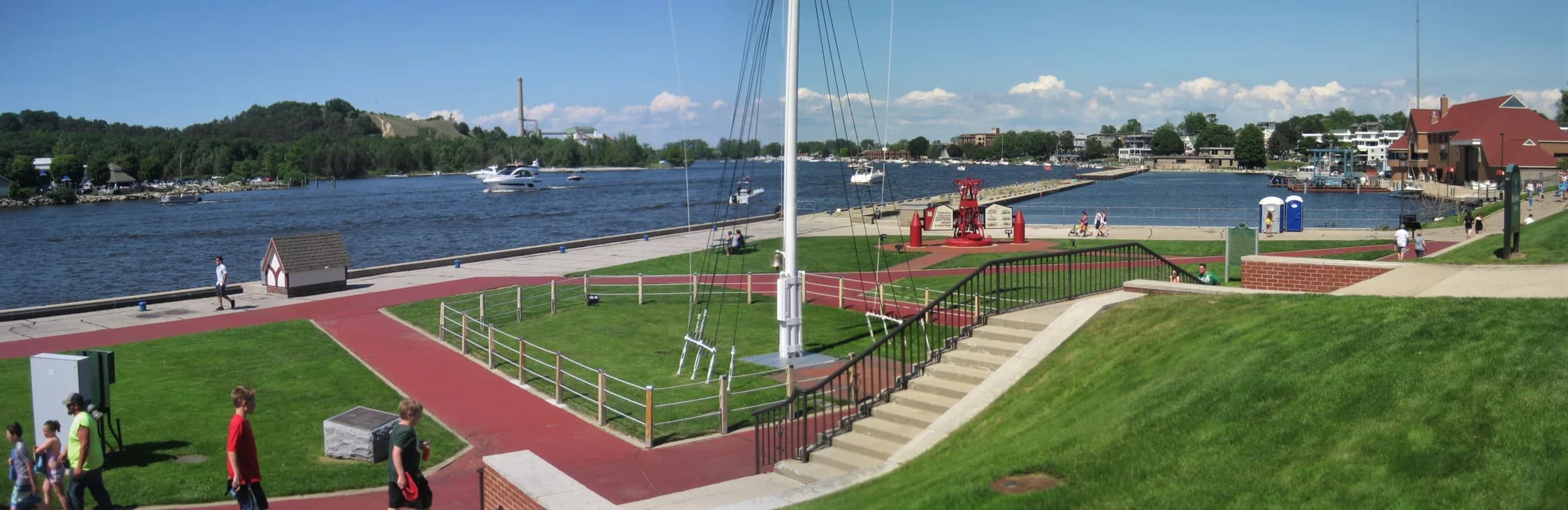 A wide panoramic view across mown parkland and a paved waterfront walk, with masts and moored boats along the far bank and low buildings behind them.