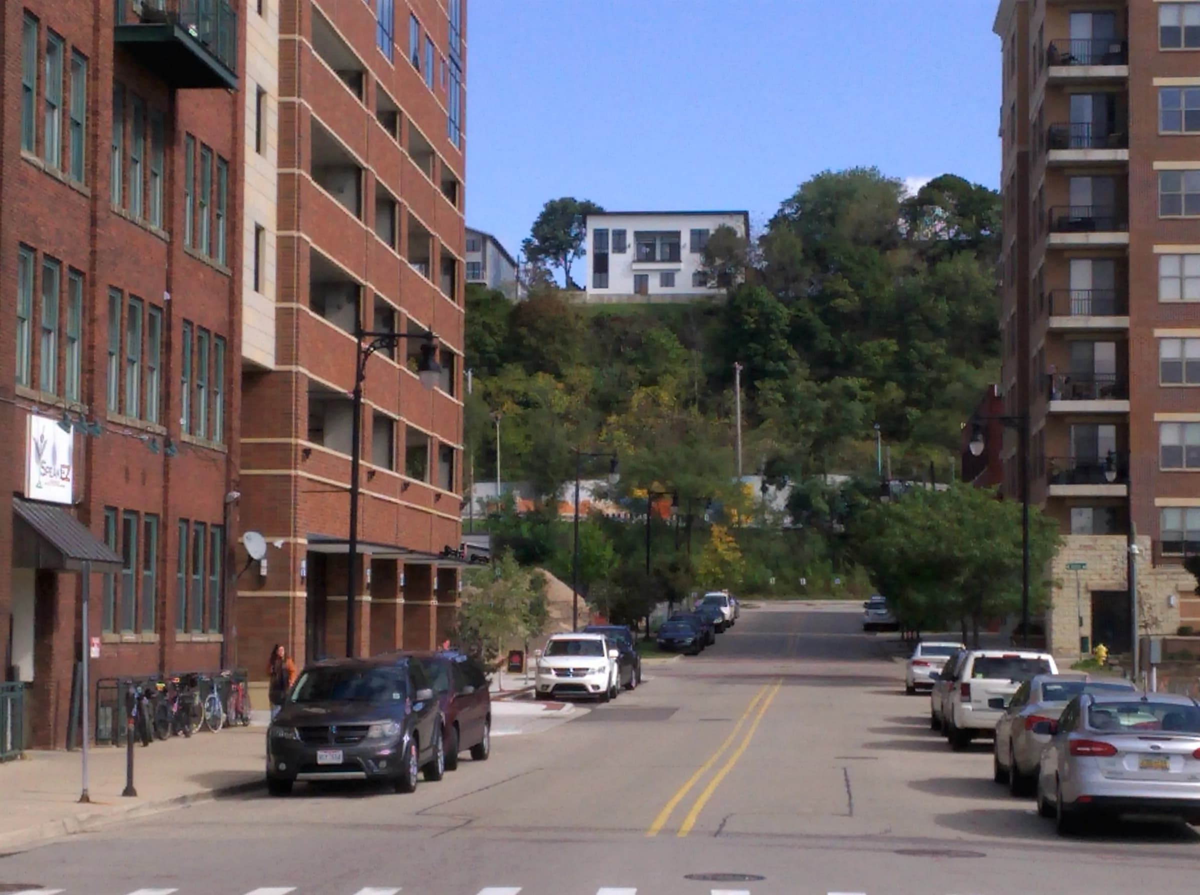 A city street climbing between mid-rise brick and concrete buildings, cars parked along both kerbs, bicycles racked at the corner and a wooded ridge rising behind the rooftops.