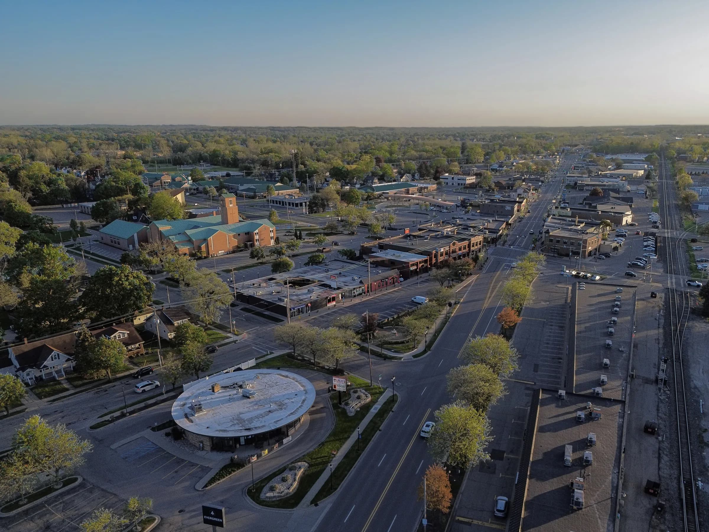 An aerial view down a wide main road lined with low shops and car parks; long evening shadows fall across the tarmac, a round flat-roofed building sits in the foreground and railway tracks run along the right side.