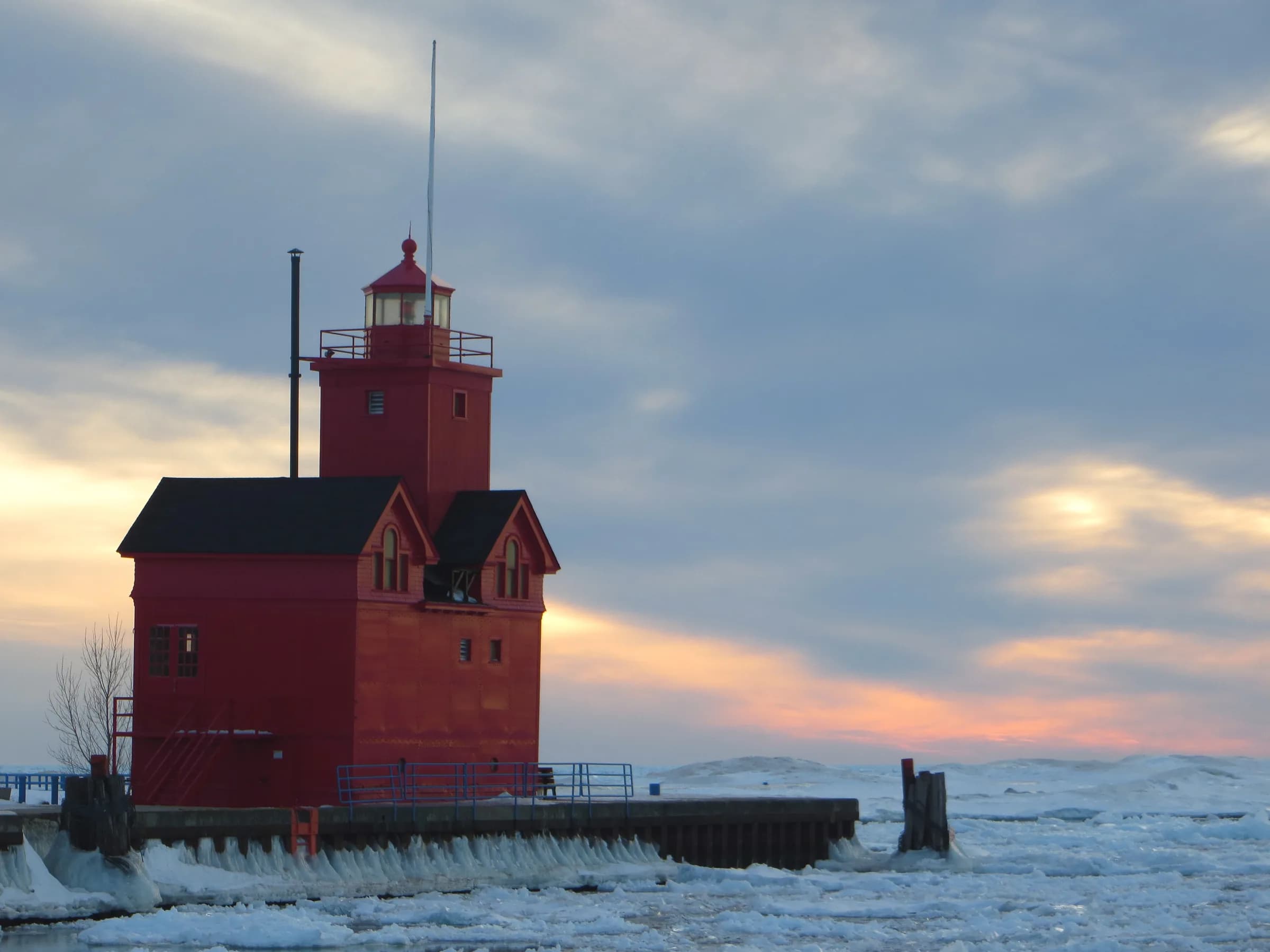 A tall red timber lighthouse with a black roof standing on a snow-dusted breakwater, lit warm by low sun against a bank of grey and gold cloud over open water.