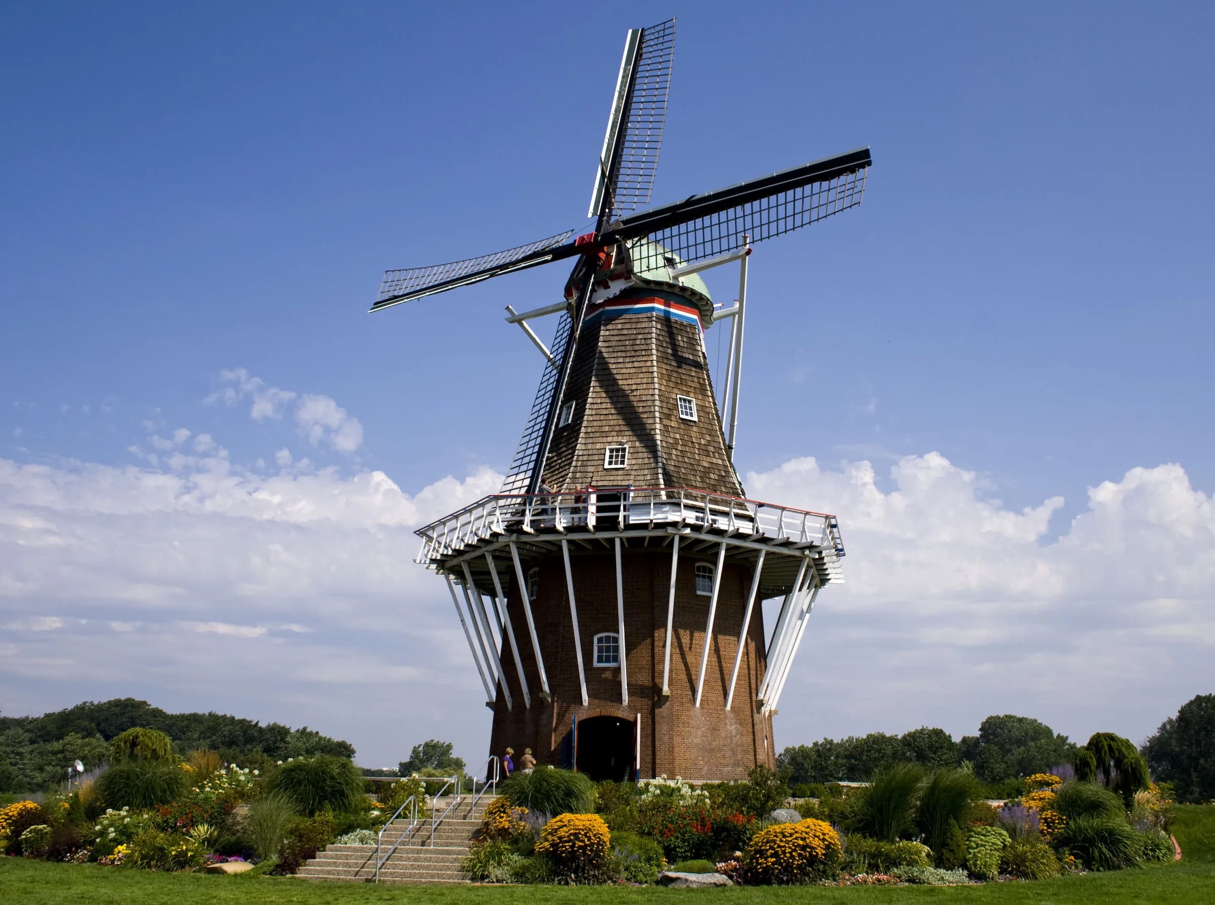 A brick and timber windmill with four full sails on a green mound, flower beds at its base, under a blue sky with scattered white cloud.
