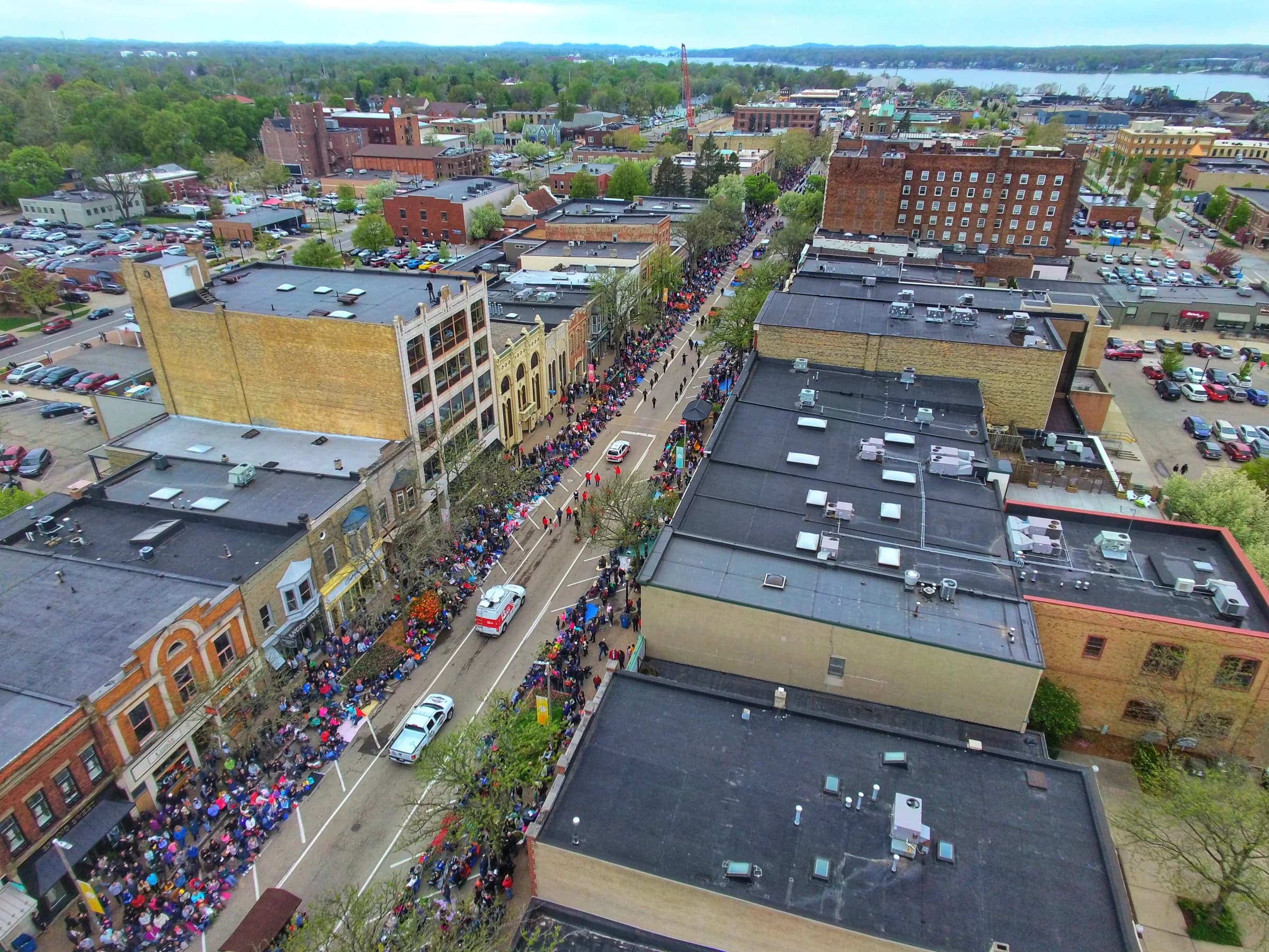 Aerial view along a downtown street lined with low brick buildings, crowds packed along both pavements and cars in the side lots, with a lake and treeline on the horizon.