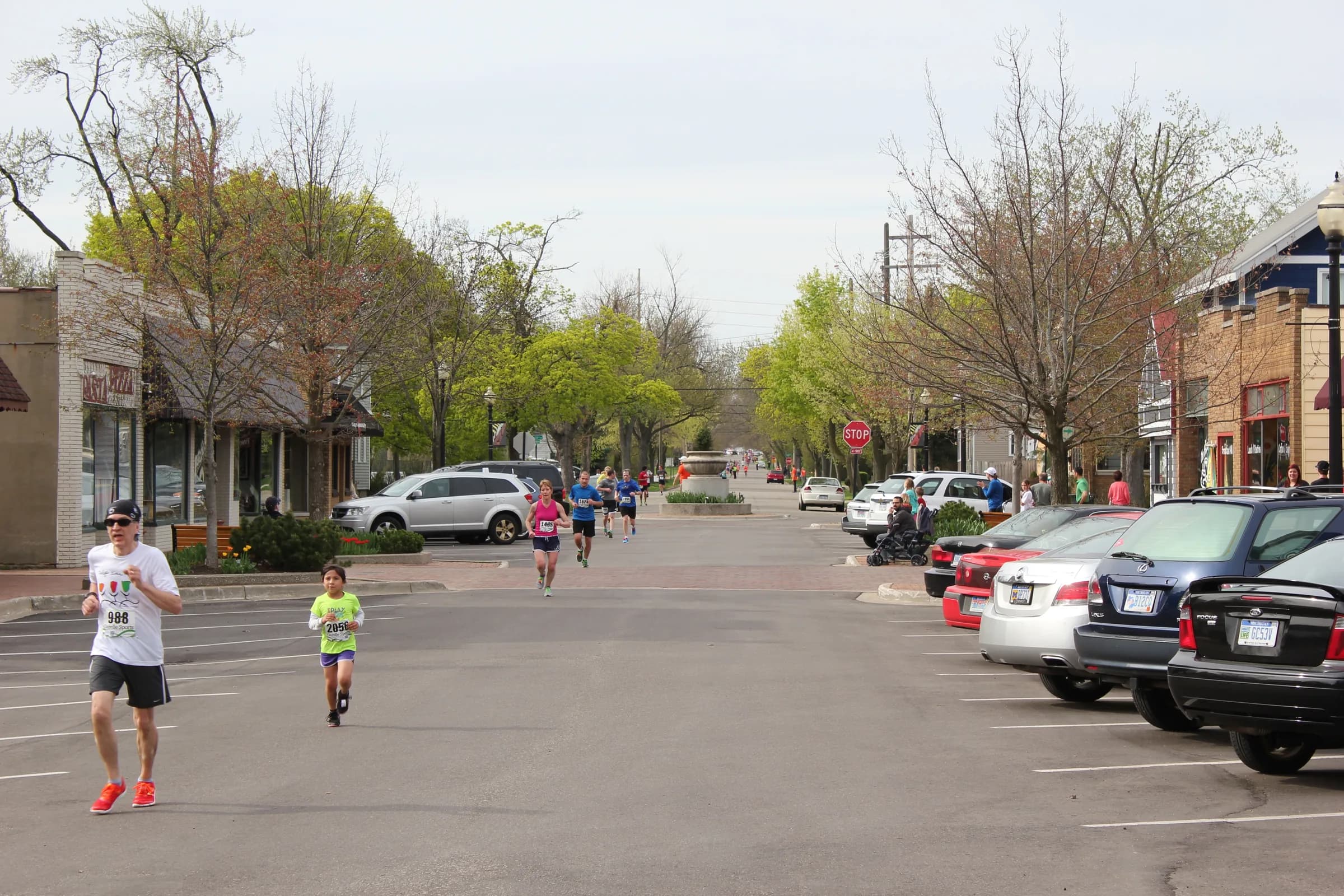 Runners in bright vests coming up a wide street between two-storey shopfronts, bare spring trees along the kerb and parked cars on both sides.