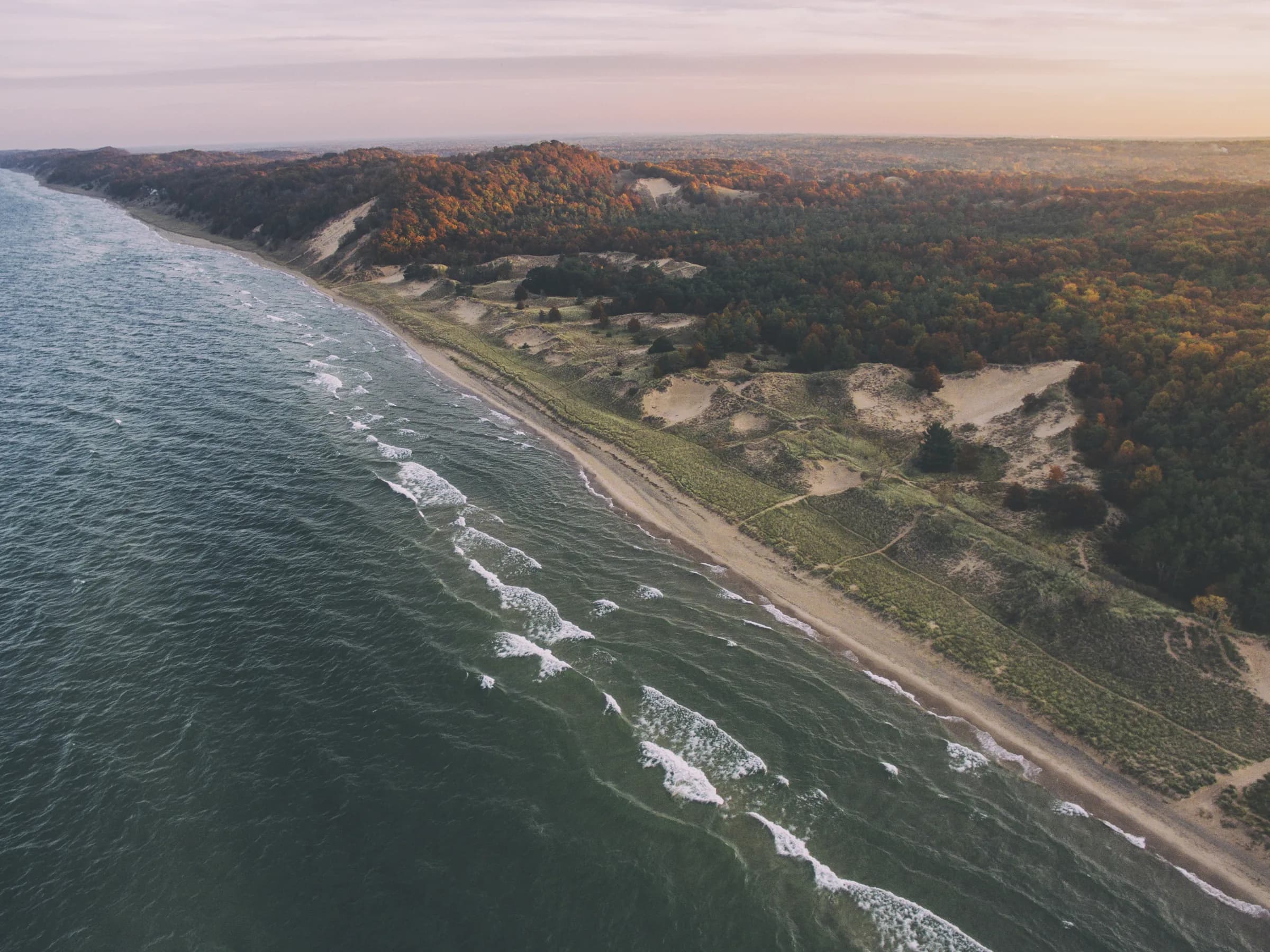 High oblique view down a long curving coastline, pale surf breaking along the sand and wooded dunes running inland under hazy afternoon light.