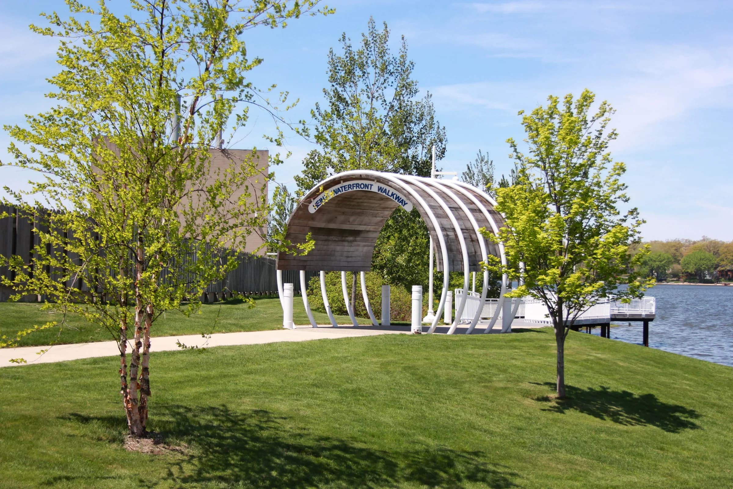 A white steel-framed pavilion with a curved roof standing on mown grass beside calm water, young trees around it and a path leading past.