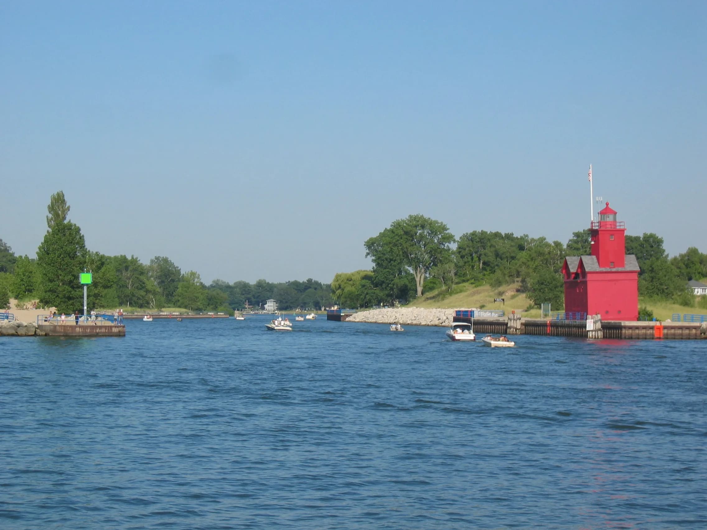 View from water level up a wide blue channel with several small boats under way, low wooded banks on both sides, a green navigation marker on the left pier and a tall red timber lighthouse standing on the right pier.
