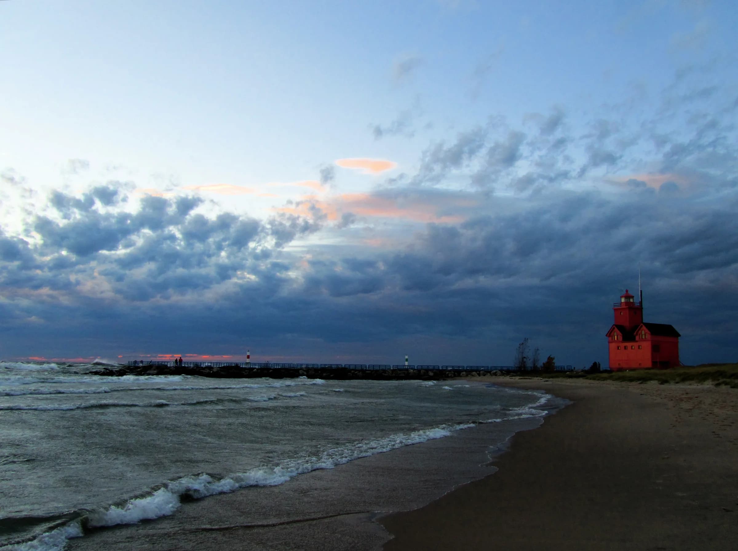 Evening on a sand beach with waves breaking in from the left, a long low breakwater running out to the horizon under heavy blue-grey cloud broken by pink light, and a small red lit building standing on the pier at right.