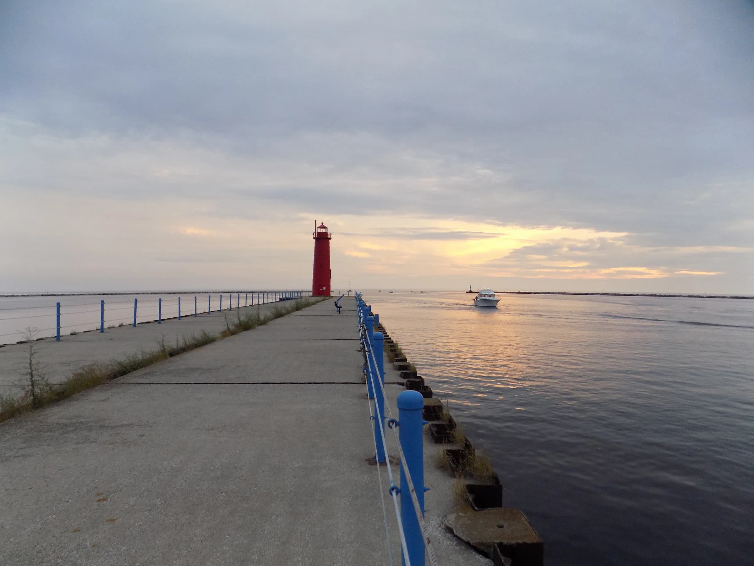 A long concrete pier with a blue steel handrail running straight out toward a small red lighthouse, low sun spreading gold across flat water and a boat on the horizon.