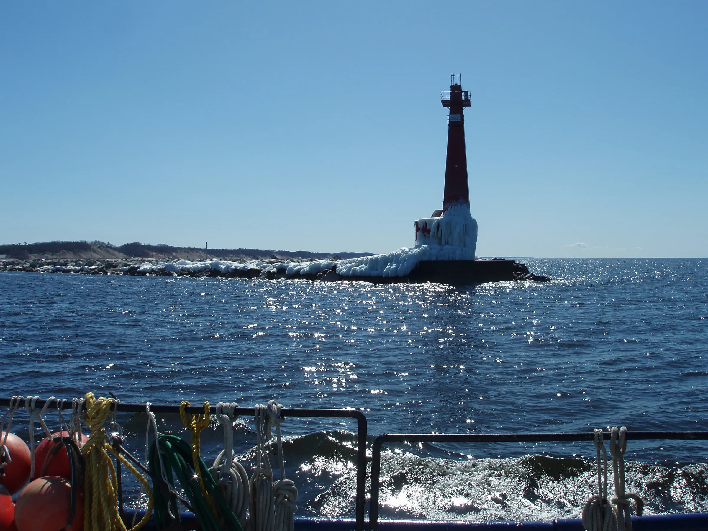 A white steel lighthouse tower on a breakwater under a hard blue sky, waves breaking white against the concrete and fishing rods propped along a railing in the foreground.