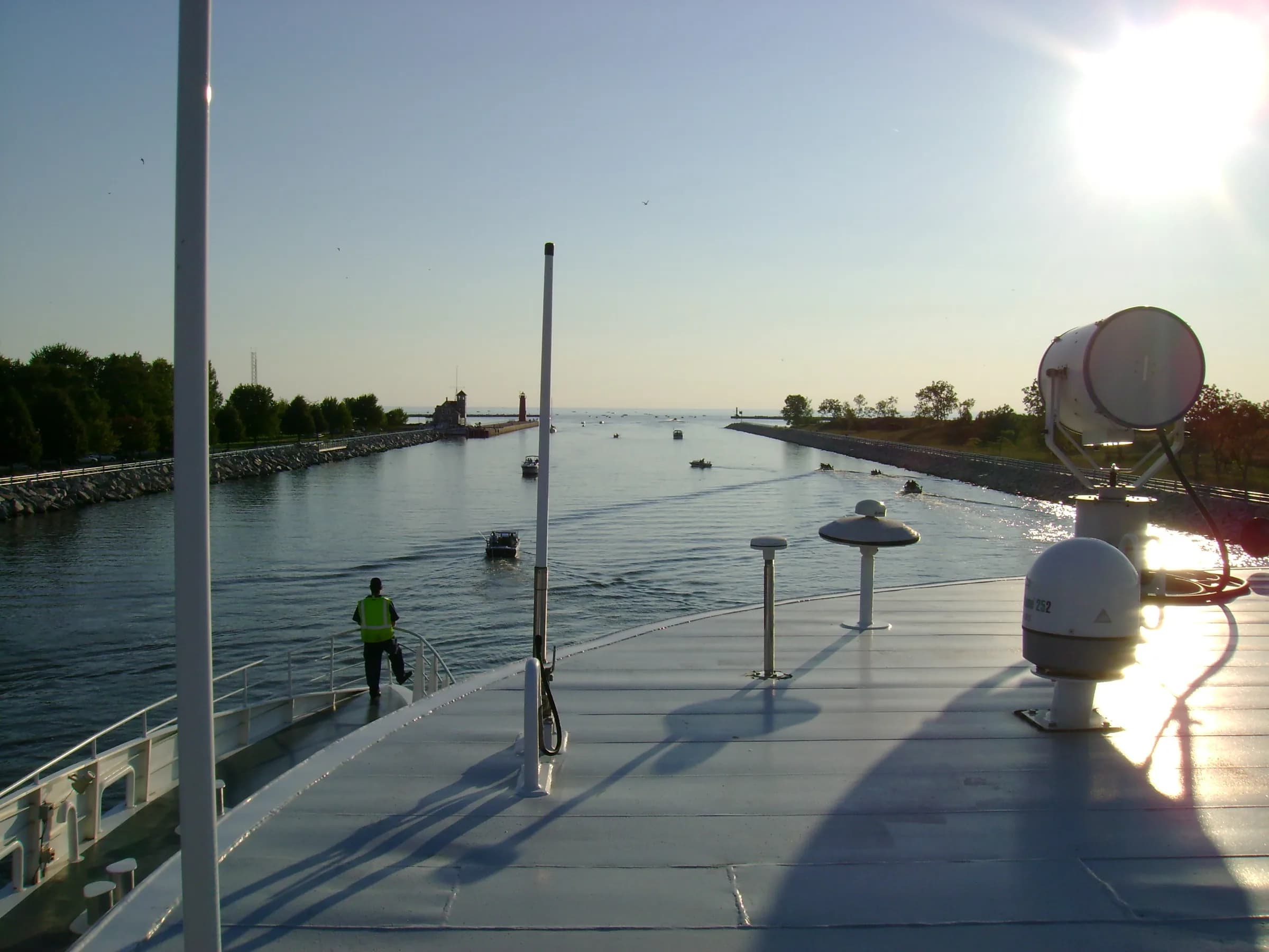 A broad straight channel seen from a walkway, lamp posts and mooring bollards receding along the right, low sun ahead and open water at the far end.
