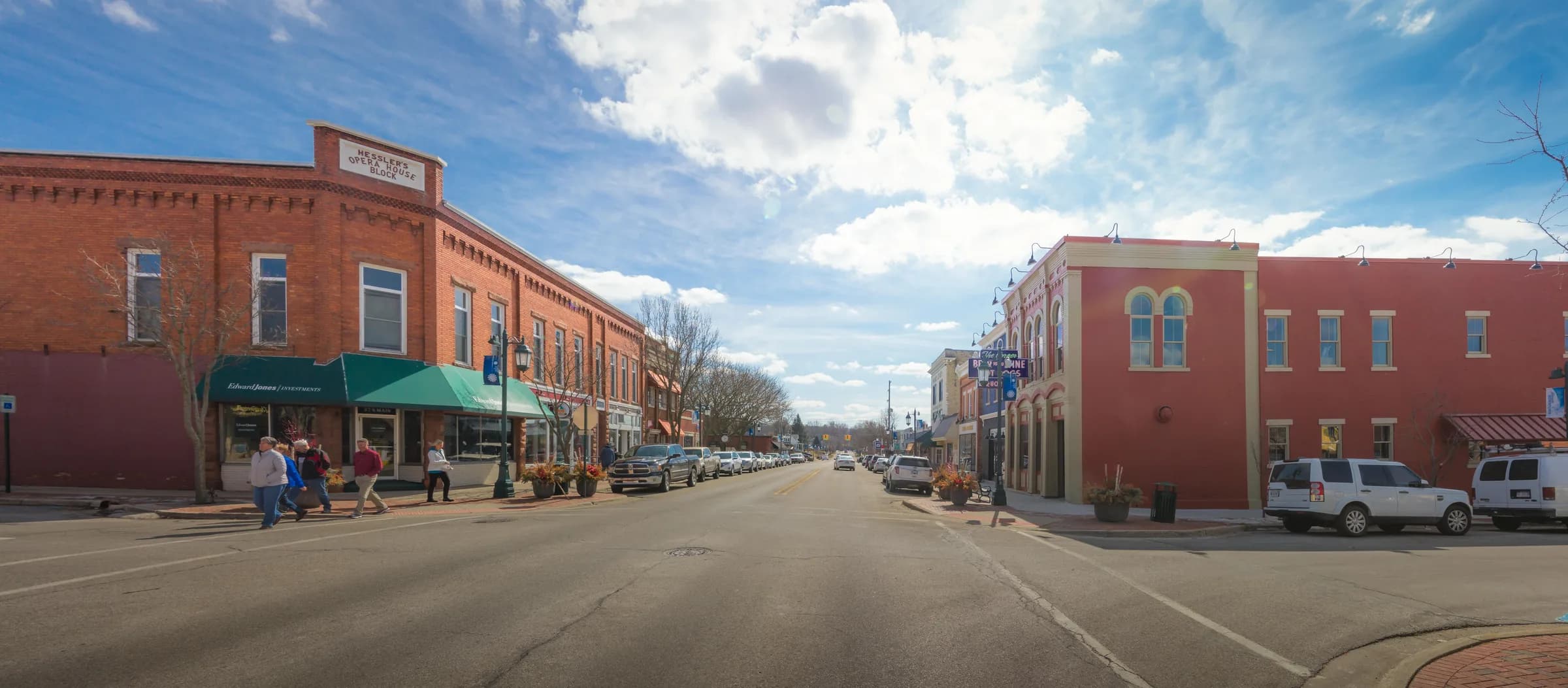 A very wide street-level view of a small-town main street: a red brick Victorian commercial block with a green awning on the left, a red-painted building on the right, cars parked along the kerb and people crossing at the corner under a bright sky.