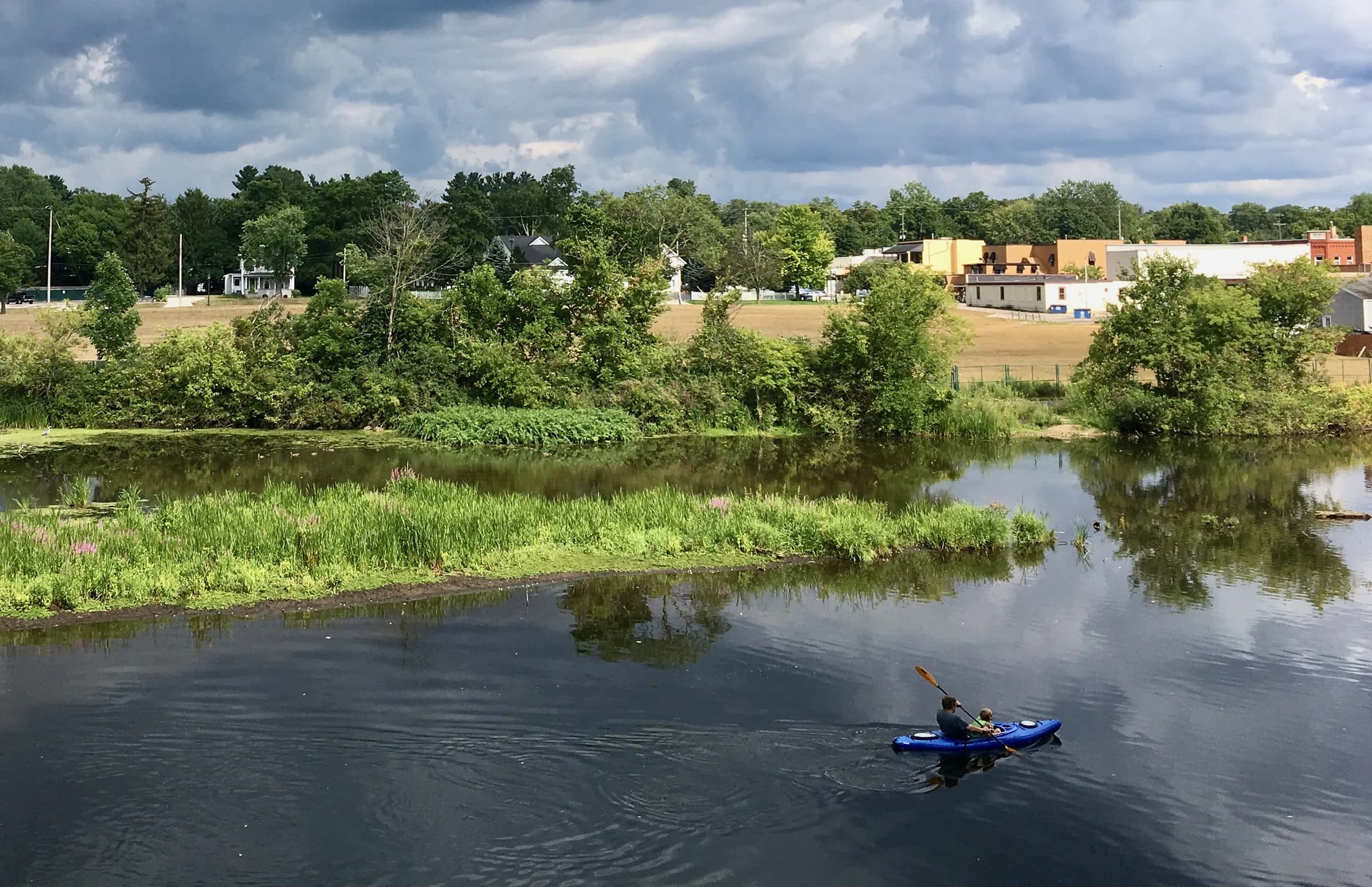 Two people in a blue kayak paddle across dark water in the lower right of the frame; a grassy island and low buildings line the far side beneath a heavy grey-blue sky.