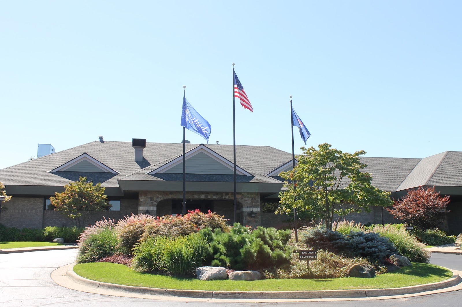 A stone and shingle clubhouse with a steep grey roof seen across a circular drive, three flagpoles at the centre of a bed of shrubs and ornamental grasses in front of it.