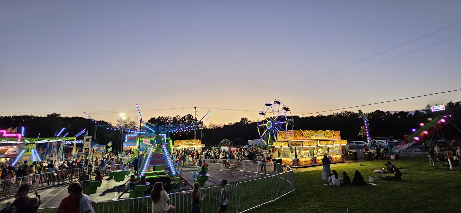 A funfair at dusk seen across an open field, lit rides and a big wheel strung along the horizon under a violet sky, with crowds silhouetted in the foreground.