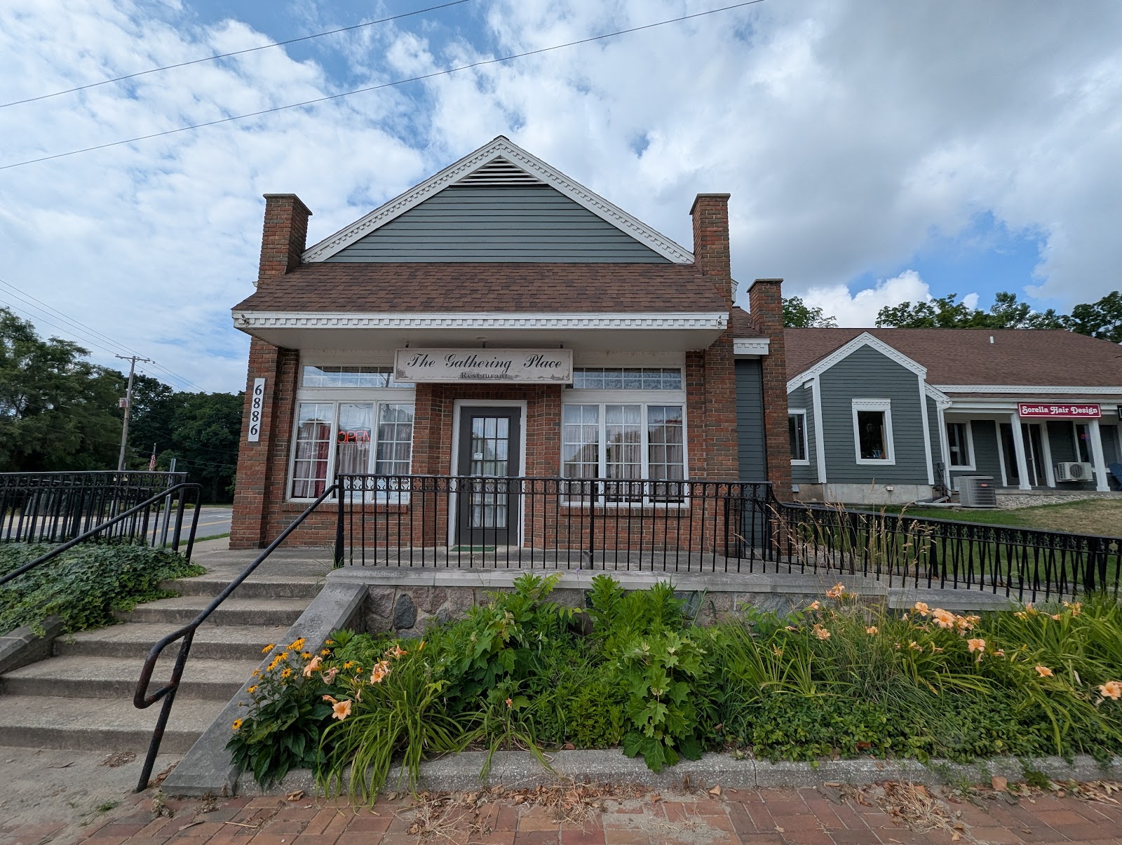A small nineteenth-century brick and clapboard commercial building with a shallow porch and iron railings, planted beds along its front and a low garden wall at the pavement.