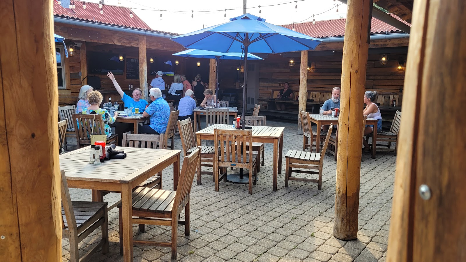 An open timber-framed terrace with tables under blue umbrellas, people eating in the late sun, and heavy log posts carrying the roof above a paved floor.