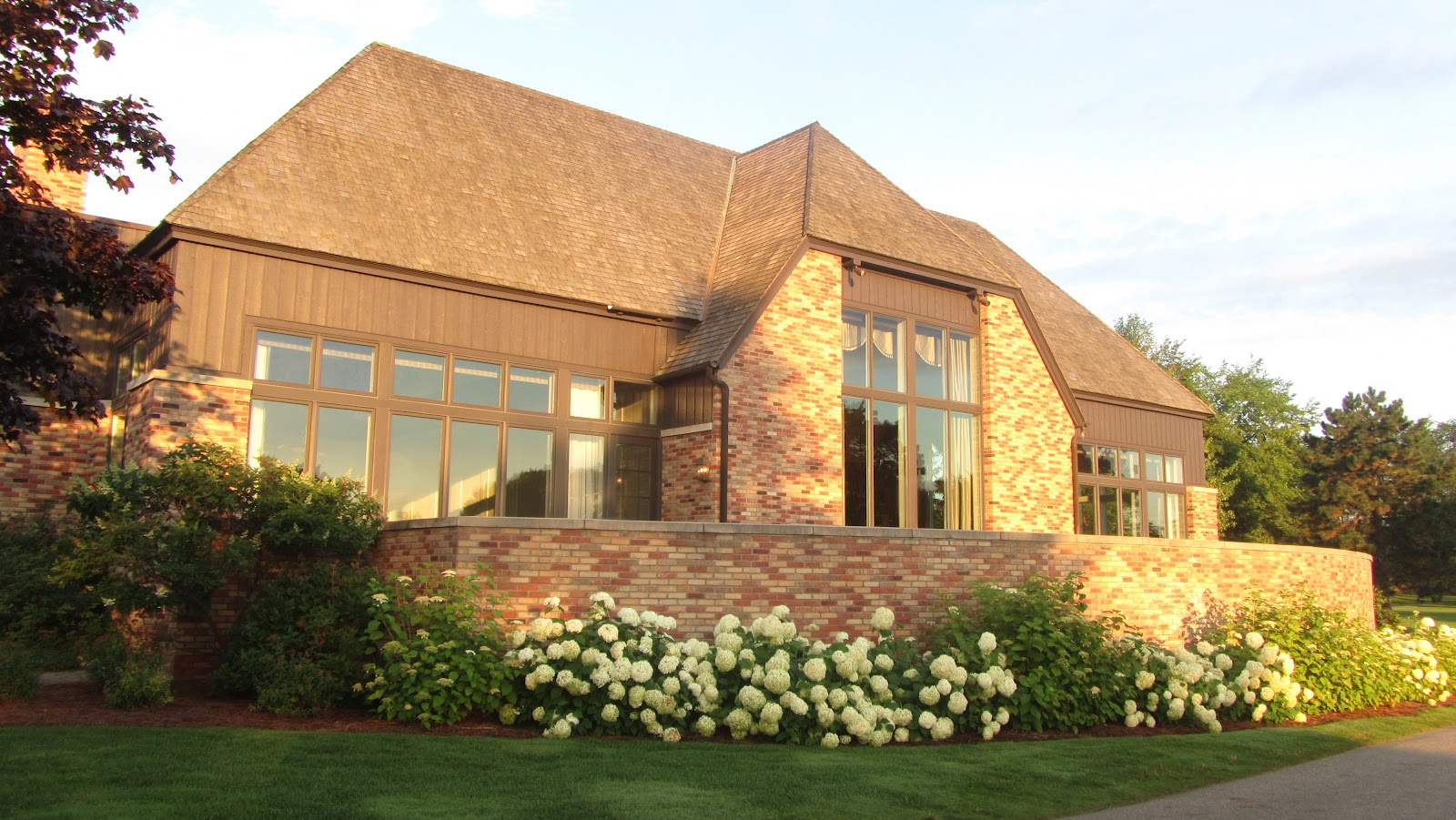 A long low stone and timber clubhouse with a steep hipped roof and tall windows lit from within, banks of white hydrangeas along the path in front of it.