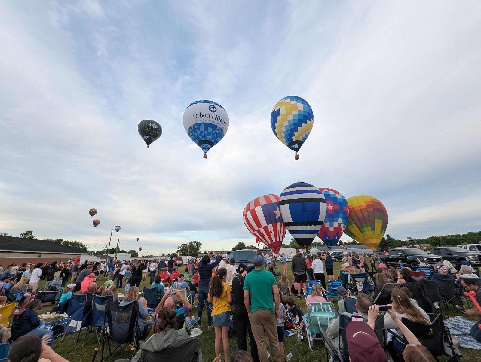 Half a dozen hot-air balloons rising into a pale cloudy sky above a crowd of people standing on grass, one balloon still being inflated at ground level.