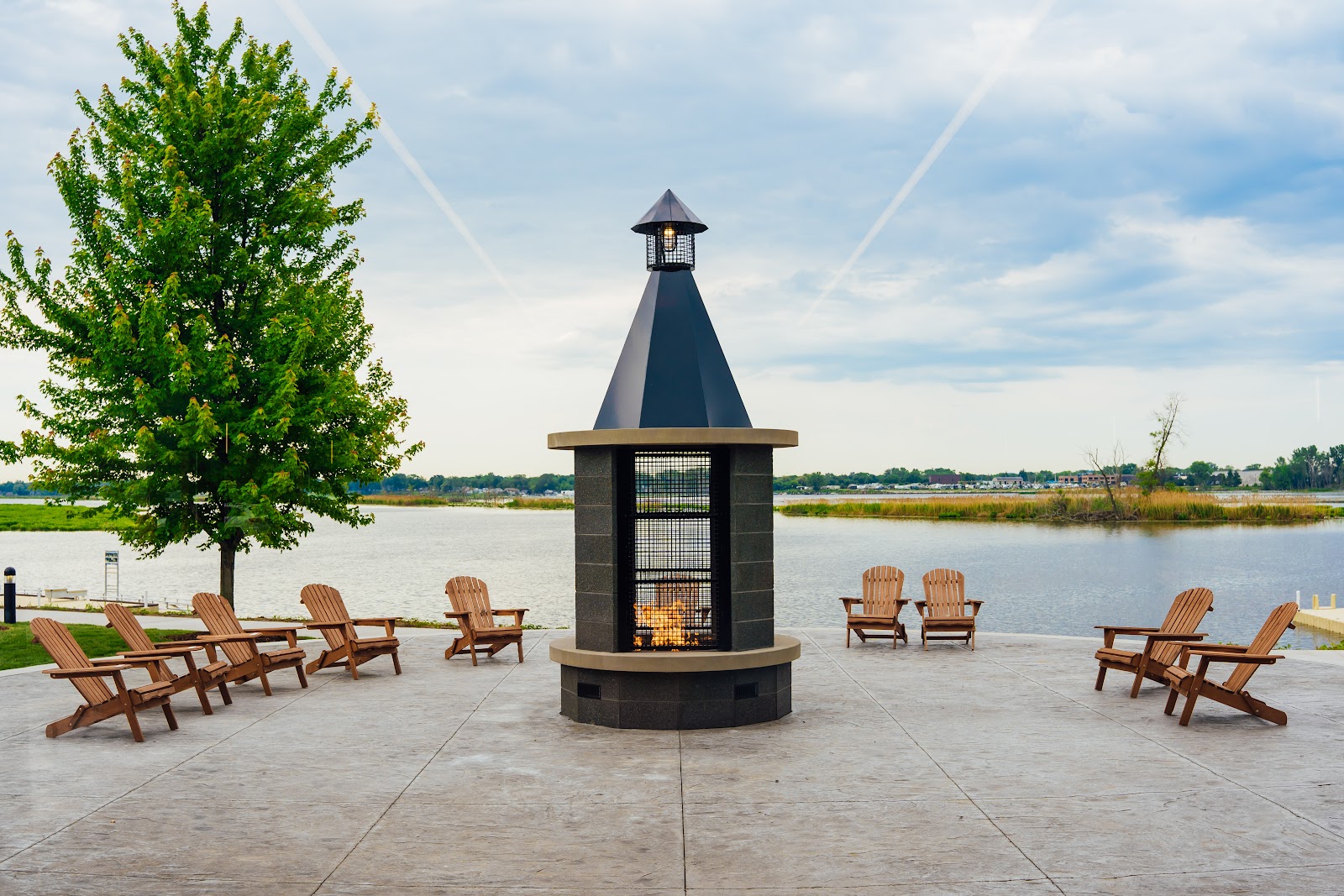 Wooden Adirondack chairs arranged in a semicircle on mown grass facing flat open water, a small dark pavilion beside them and young trees either side.