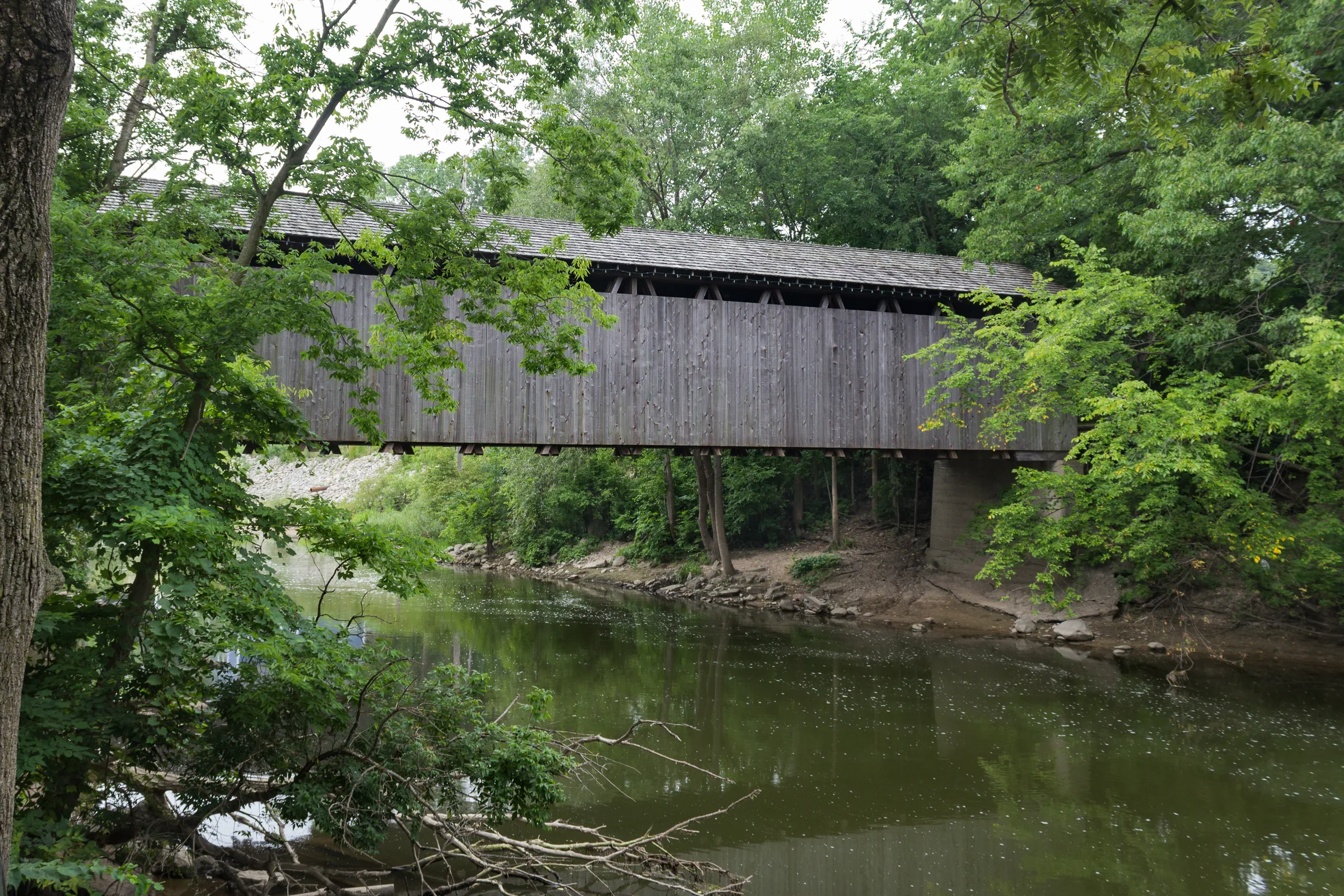 A long timber bridge with weathered grey siding spans slow green water, framed by heavy summer foliage, with fallen branches and a soft reflection in the river below.