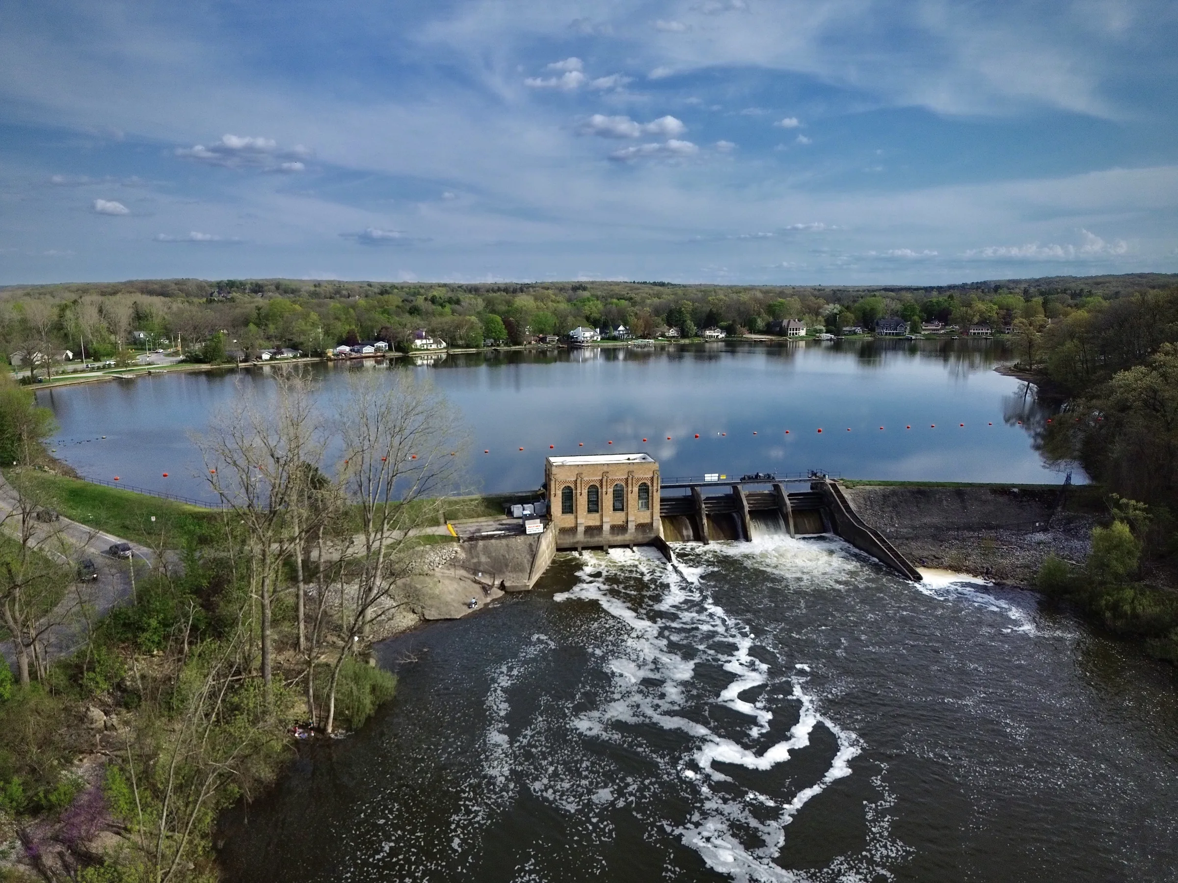 An elevated view of a low concrete dam and a small brick powerhouse; white water spills across the full width of the frame while a wide calm pond and wooded banks lie beyond under a blue sky with scattered cloud.