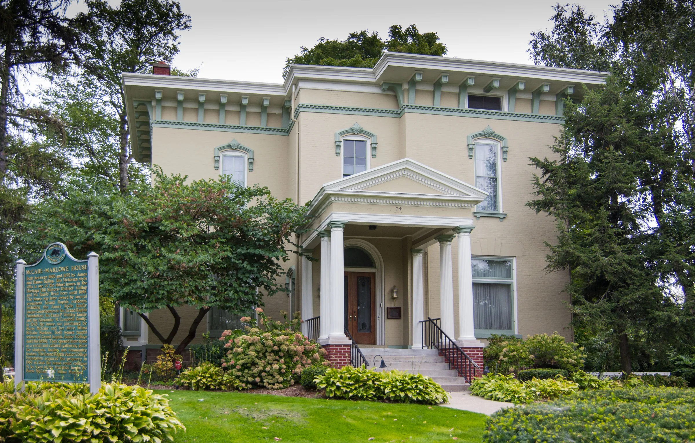 A two-storey cream-rendered house with tall arched windows, deep bracketed eaves and a small columned entrance porch reached by brick steps, framed by mature conifers with flowering shrubs and a clipped lawn in front.