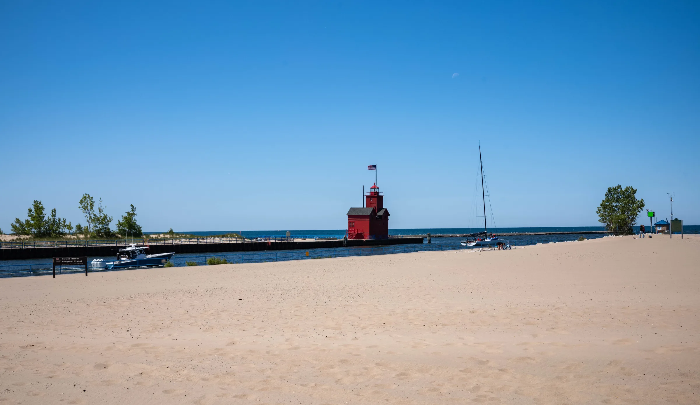 A wide sweep of pale dry sand under a deep blue summer sky, with a low breakwater and pier running out to flat open water, a moored sailboat with its mast up, and a small red structure at the far end of the pier.