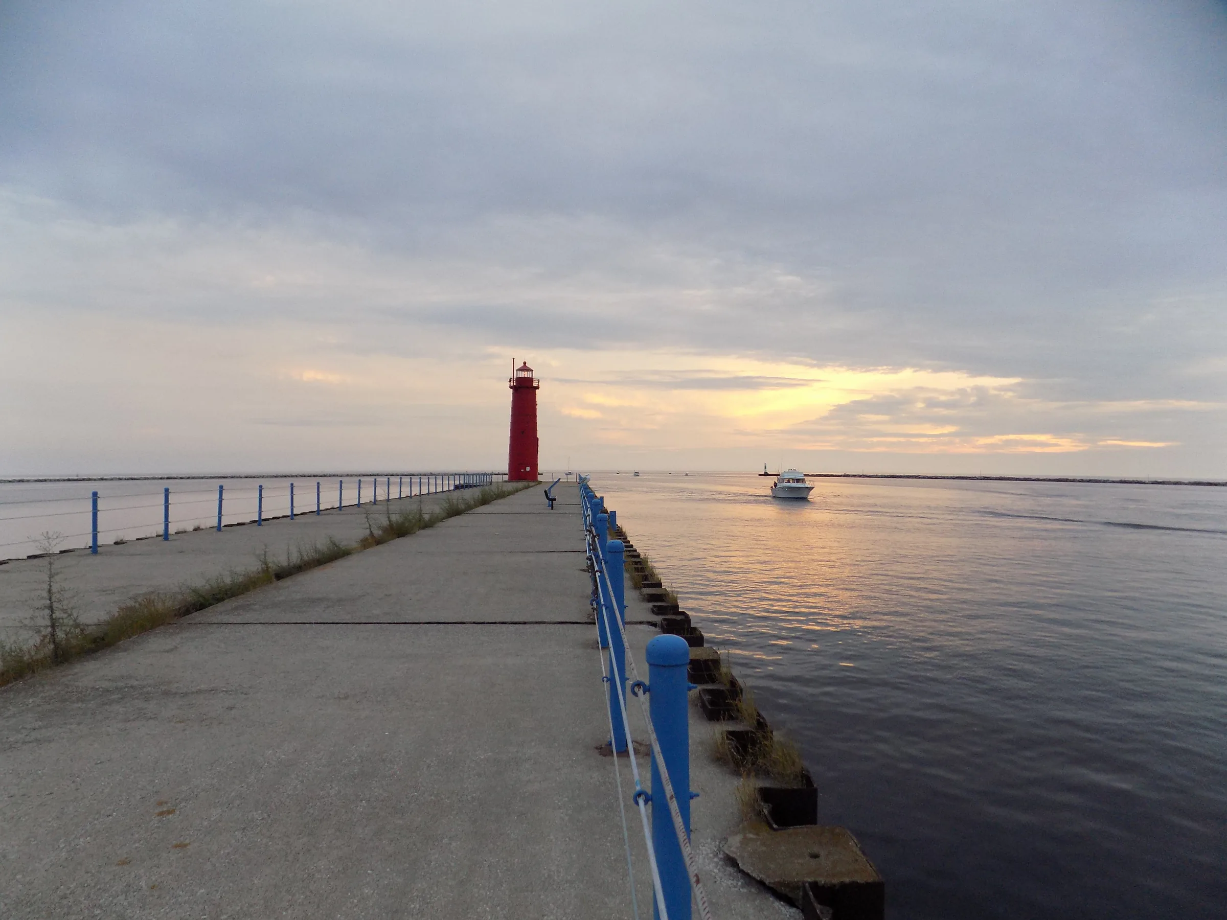 A long concrete pier with a blue steel handrail running straight out toward a small red lighthouse, low sun spreading gold across flat water and a boat on the horizon.
