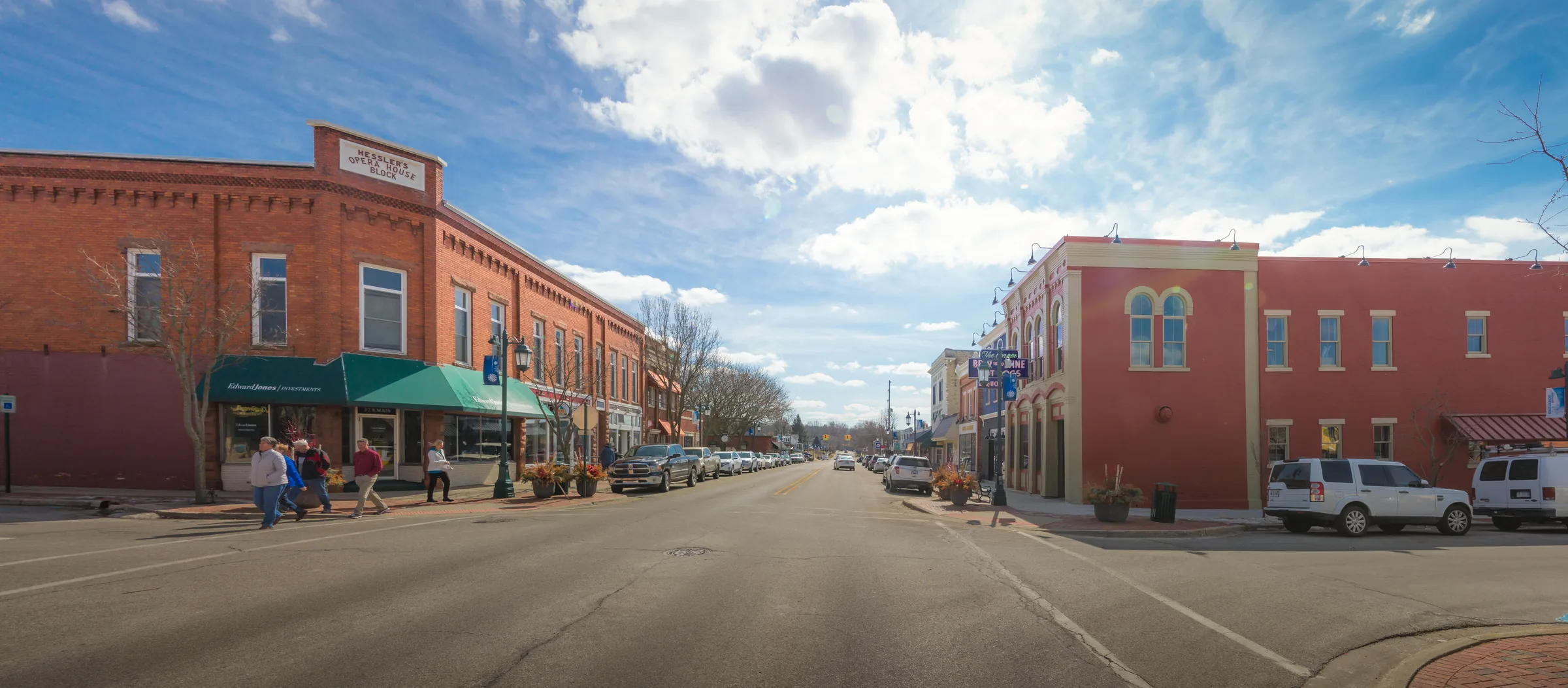 A very wide street-level view of a small-town main street: a red brick Victorian commercial block with a green awning on the left, a red-painted building on the right, cars parked along the kerb and people crossing at the corner under a bright sky.