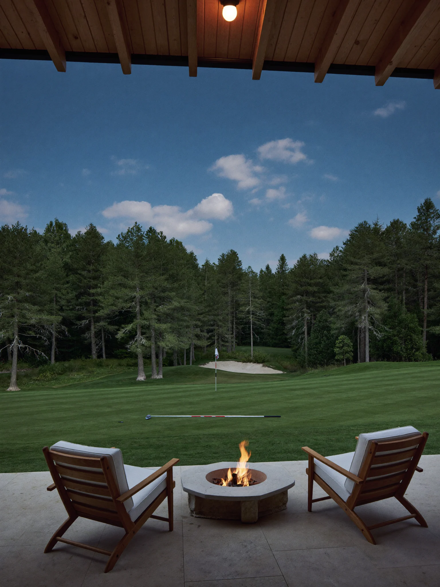 Two teak chairs and a lit fire table on a clubhouse terrace, looking across a striped fairway to a pine-backed green